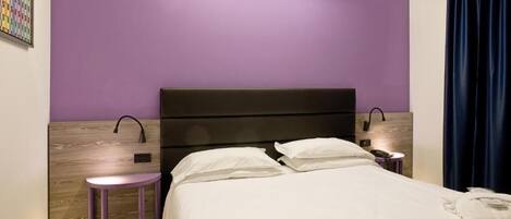Quarto de hotel com cama, mesa de cabeceira, um banquinho pequeno roxo e luminária fixada na parede.