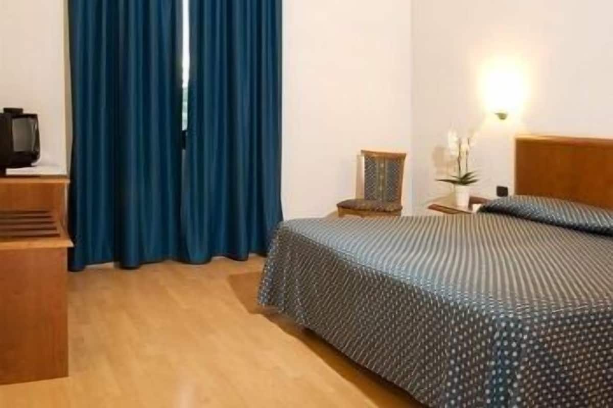 A hotel room with a bed, a wooden nightstand, a television, a chair, and a window with blue curtains.