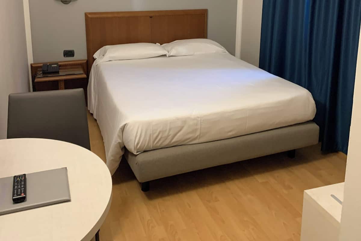 A hotel room with a bed, a bedside table with a lamp, a desk with a laptop, and a chair.