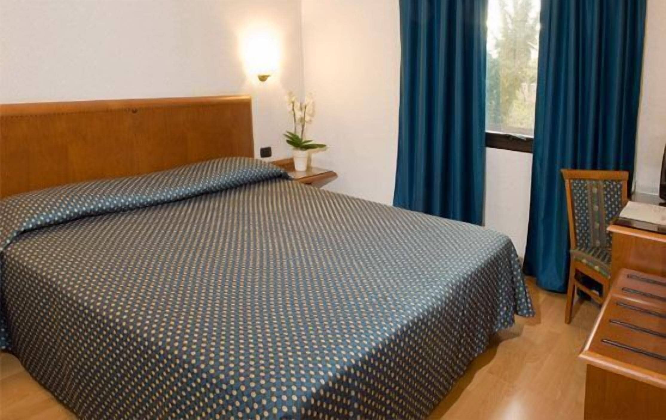 Habitación de hotel con una cama grande, una mesita de noche con lámpara, una silla y una ventana con cortinas.