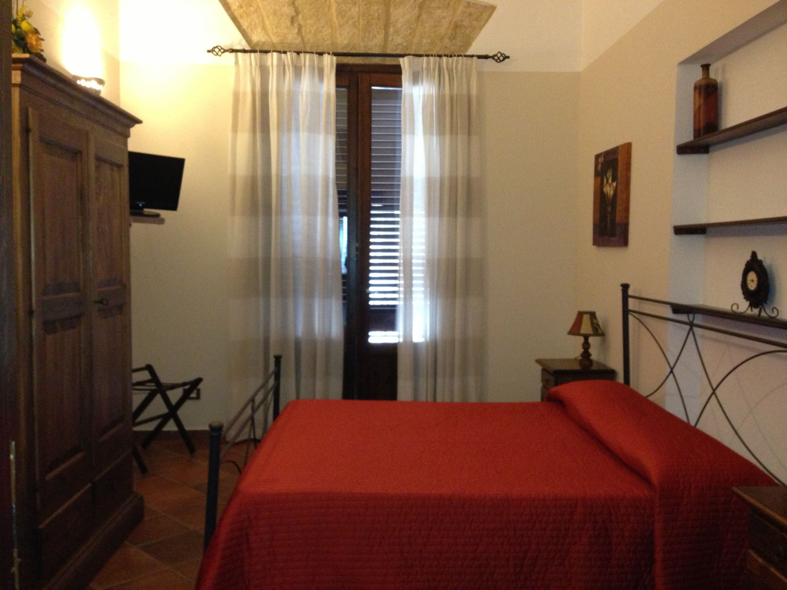 A bedroom with a bed, a wooden wardrobe, a television, a window with curtains, and a small table with a lamp.