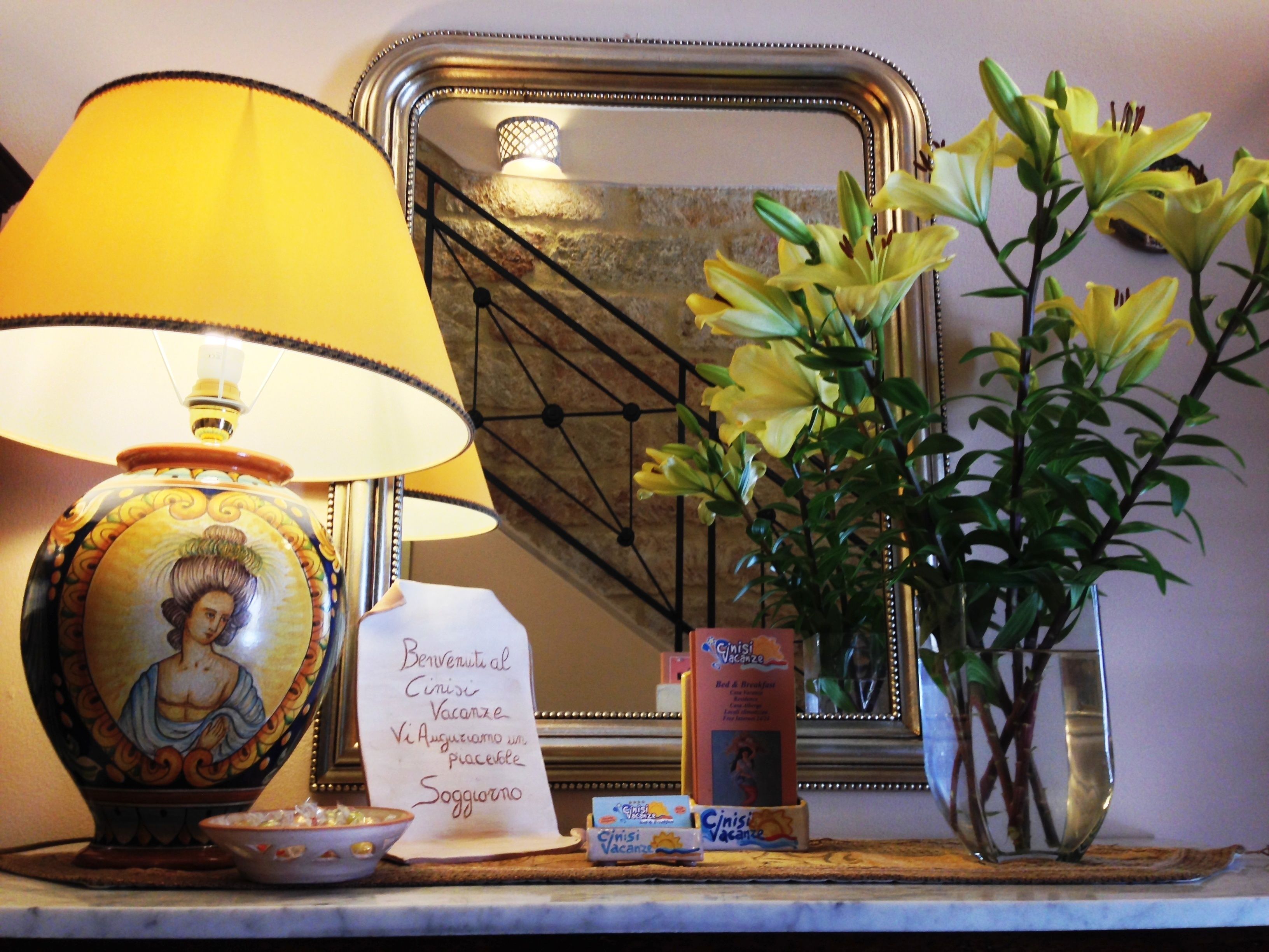 Une table avec une lampe, un miroir encadré, un vase de fleurs et un mot écrit à la main.