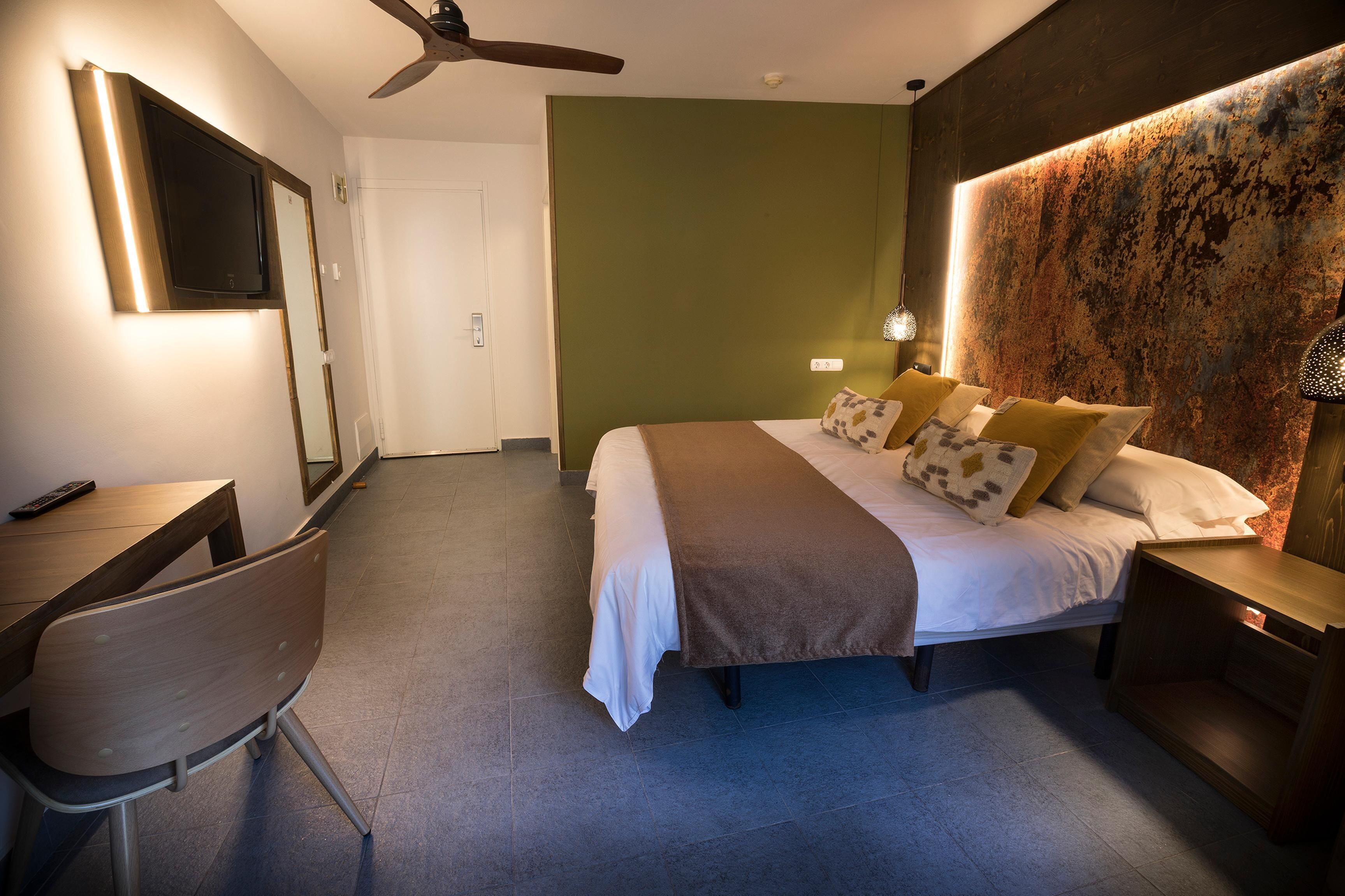A hotel room with a bed, bedside table, chair, TV, and ceiling fan.