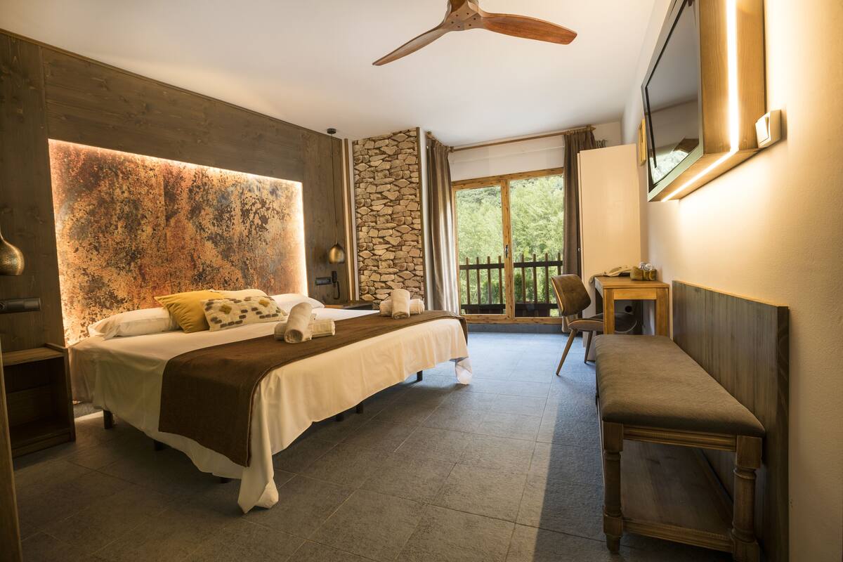 A bedroom with a large bed, a bench, a desk, a stone wall, and a balcony with a view of trees.