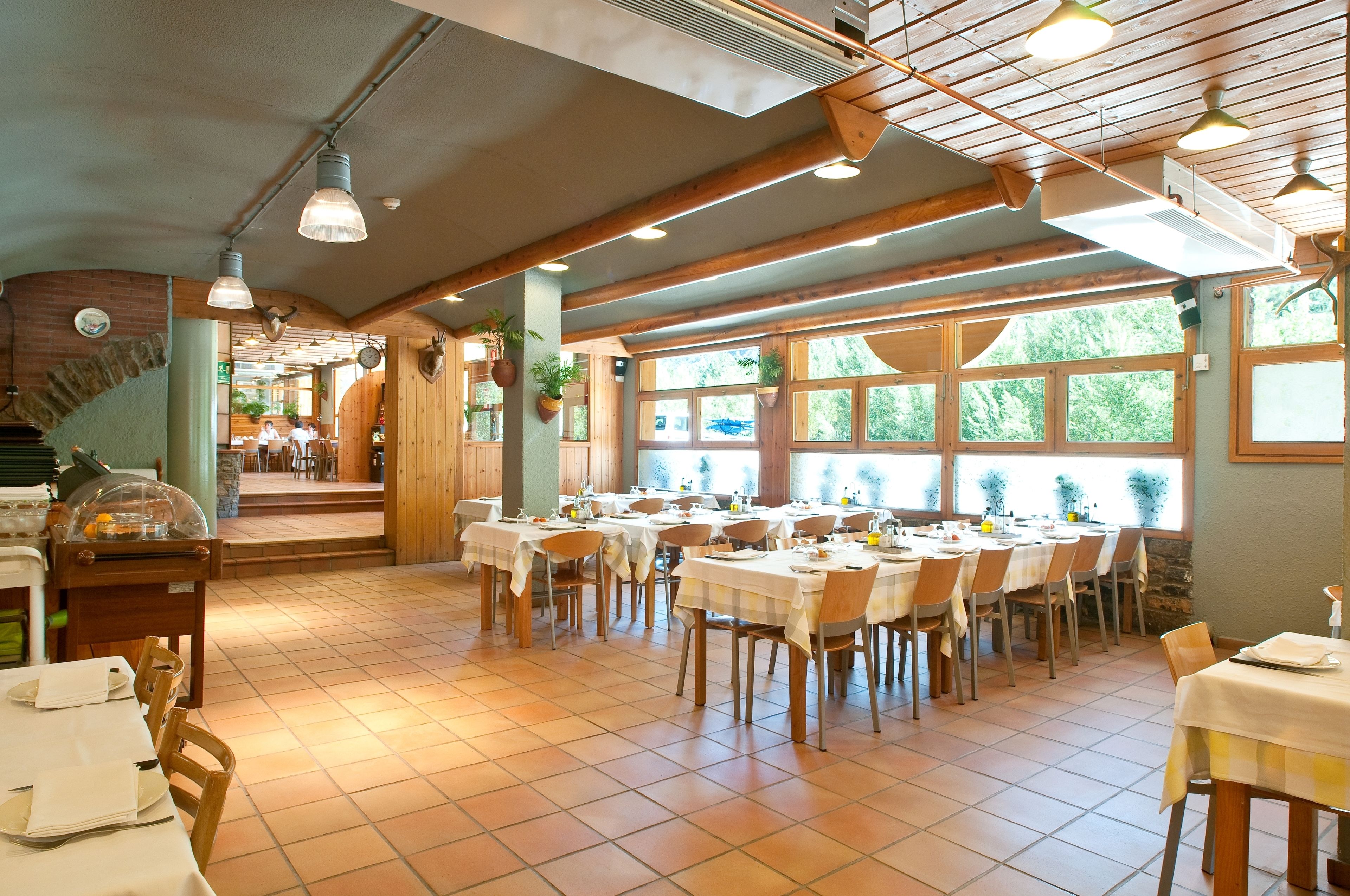 A restaurant with multiple tables and chairs, wooden beams on the ceiling, and large windows with a view of greenery outside.