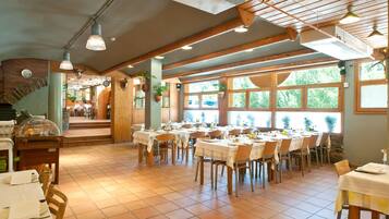 A restaurant with multiple tables and chairs, wooden beams on the ceiling, and large windows with a view of greenery outside.