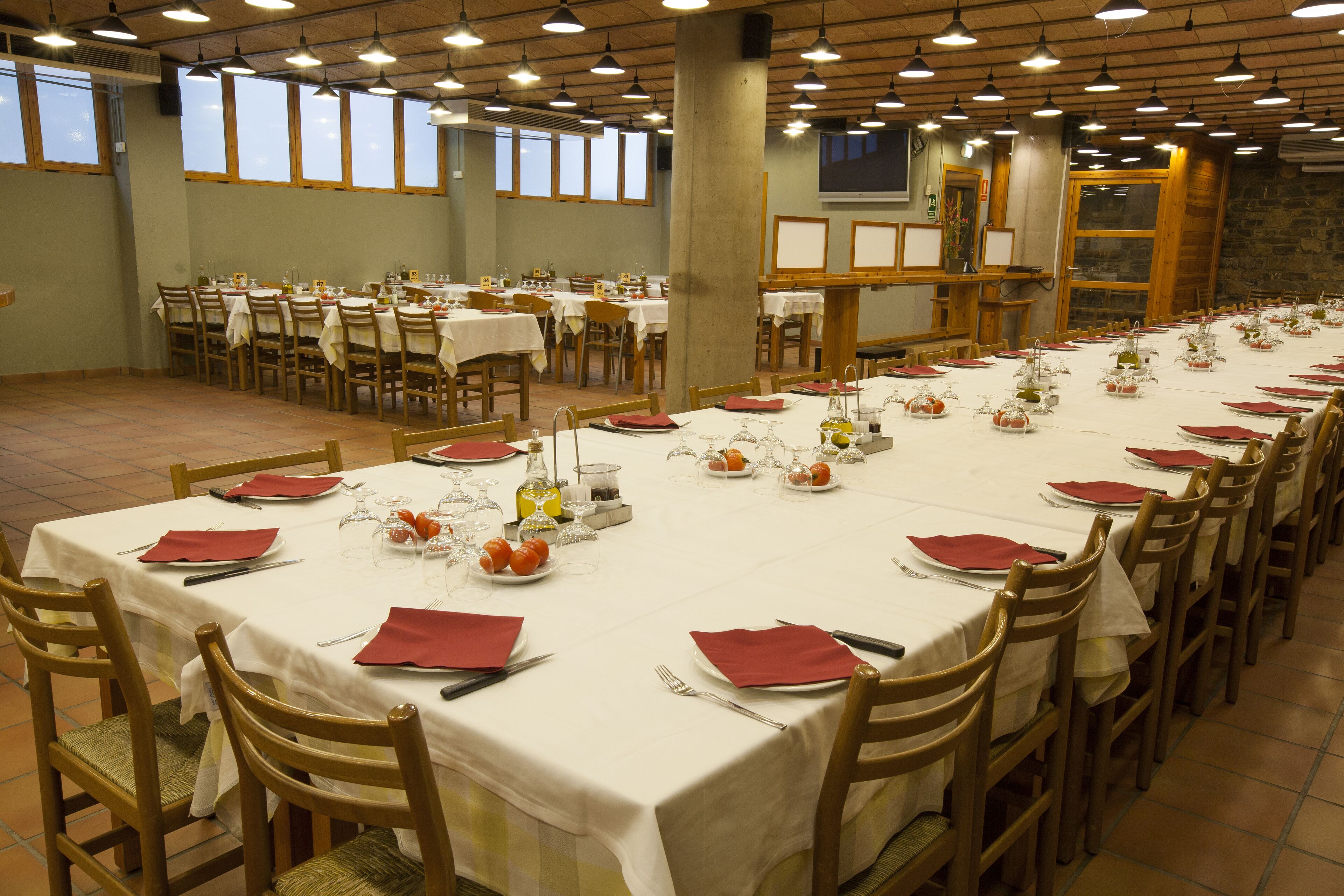 Un amplio comedor con varias mesas dispuestas para comidas, sillas de madera y una larga barra con un televisor.