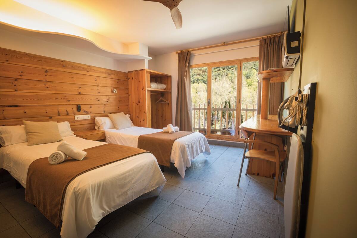 A room with two beds, a wooden headboard, a desk with a chair, and a window with curtains.