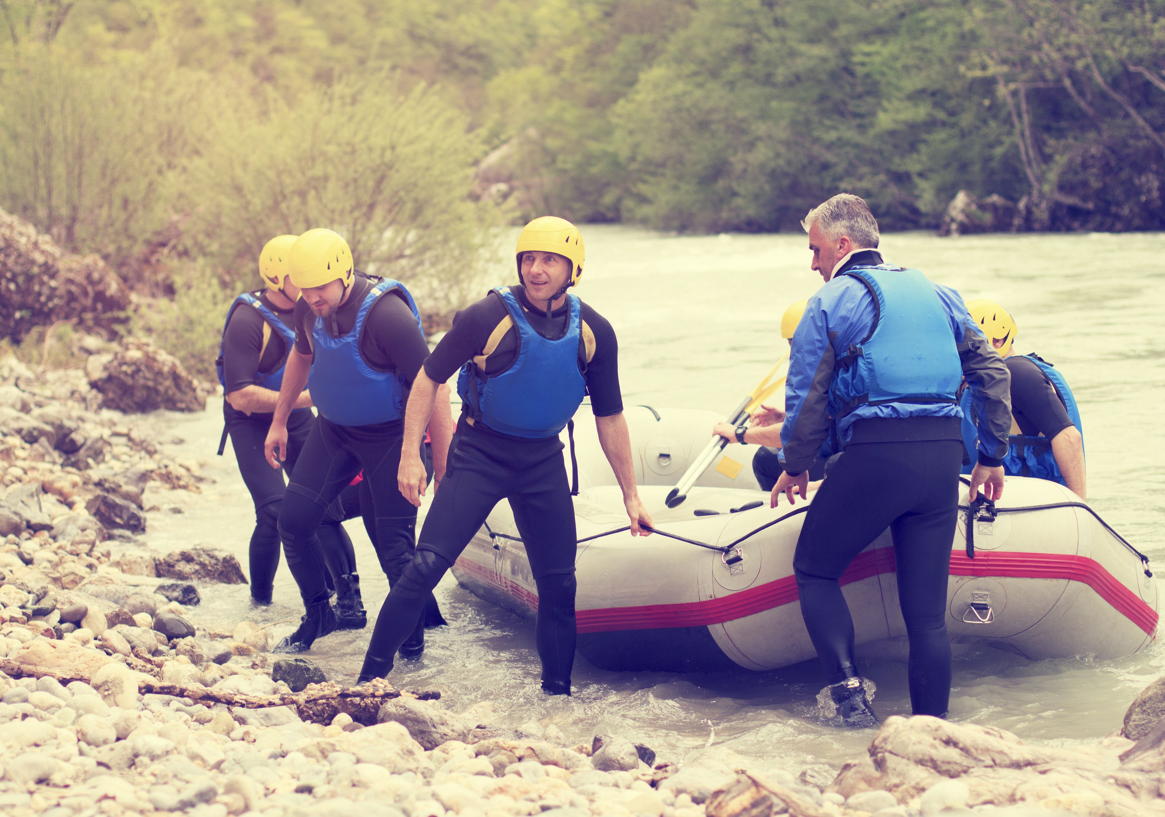 A group of people wearing life jackets and helmets, preparing to enter a white inflatable raft on a rocky riverbank.