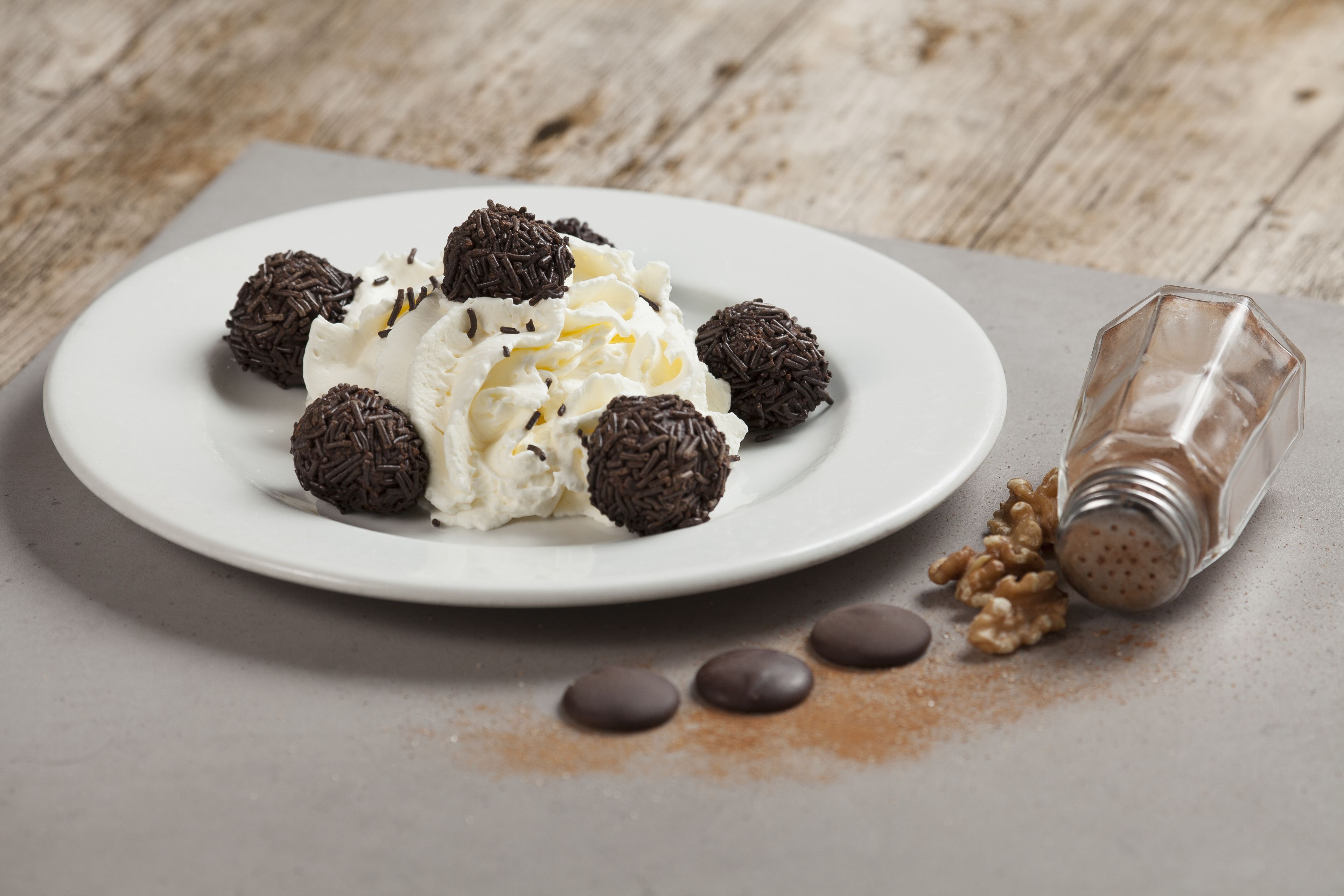 A plate of whipped cream with chocolate balls and a salt shaker on a wooden table.