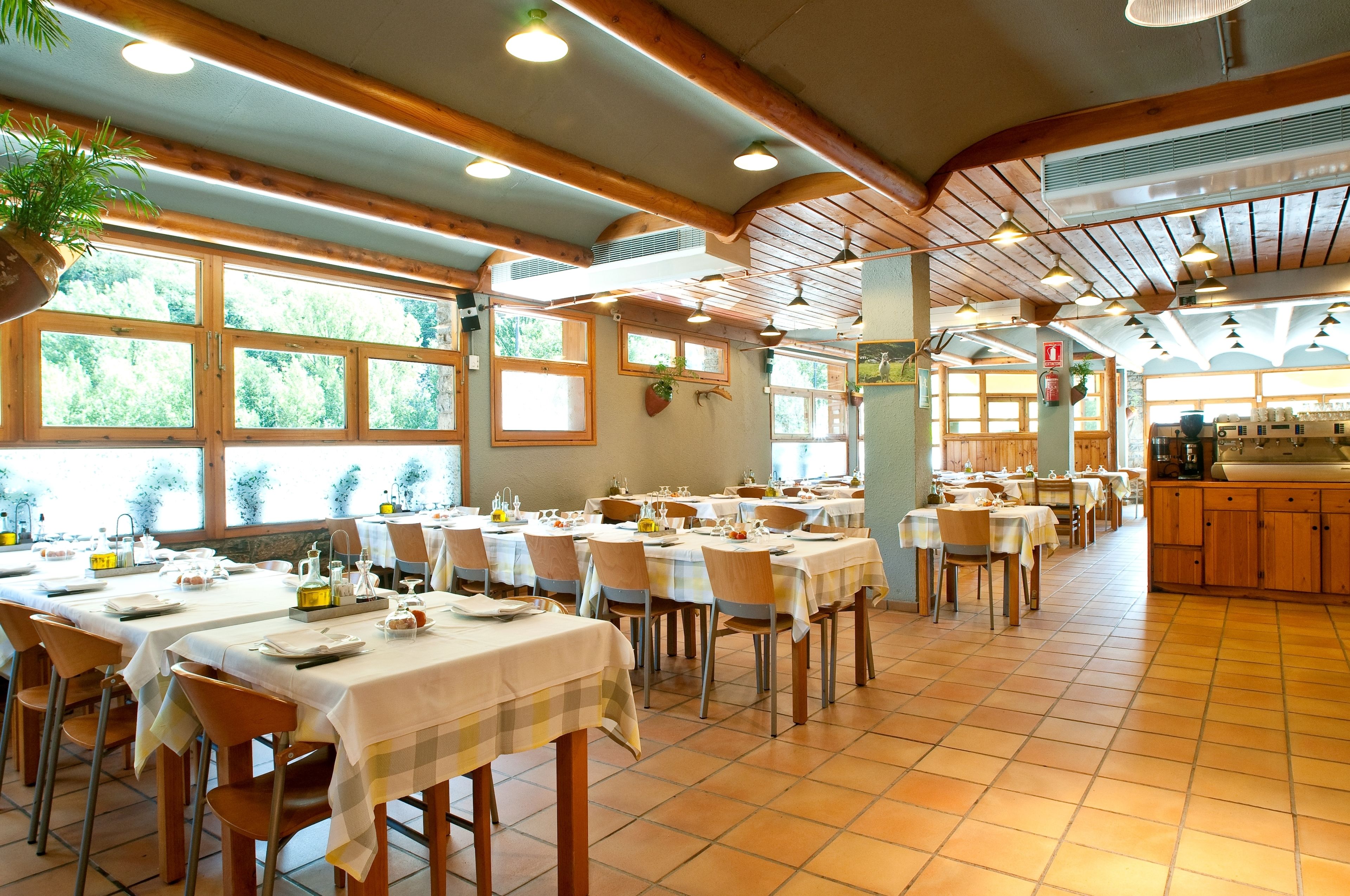 A dining area with multiple tables set for service, wooden beams on the ceiling, and large windows with a view of trees.