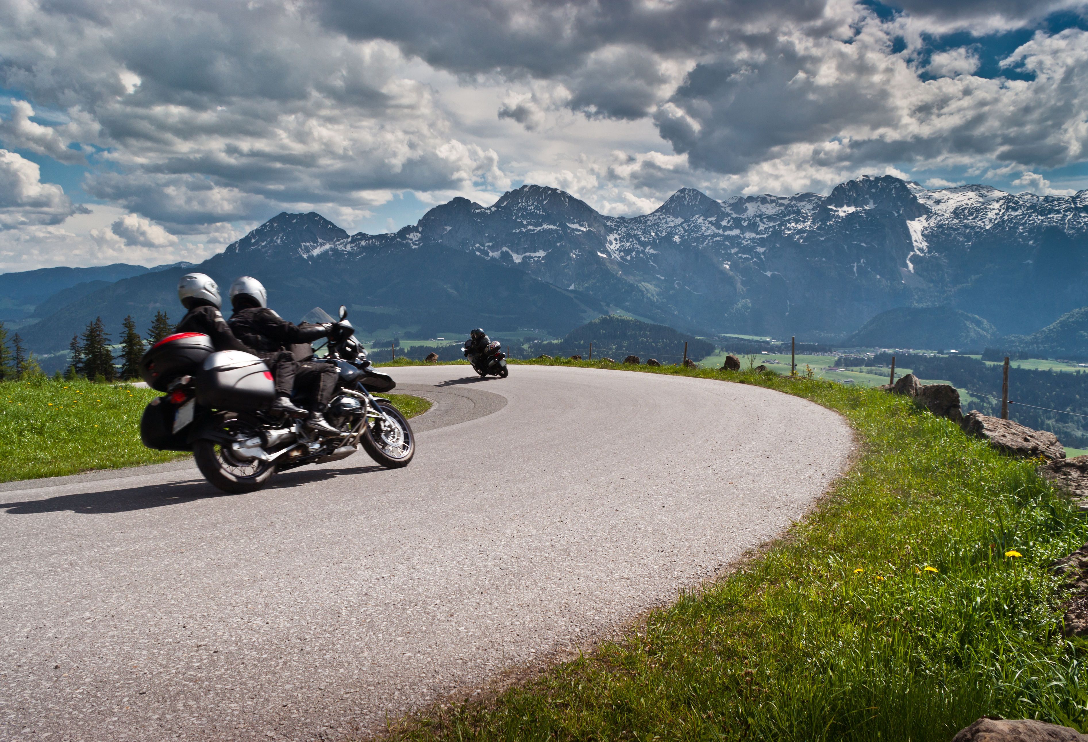 Zwei Motorradfahrer auf einer kurvenreichen Bergstraße, im Hintergrund schneebedeckte Gipfel.