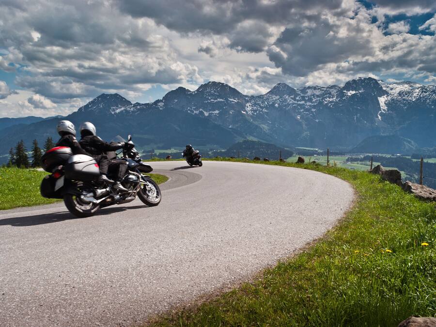 Zwei Motorradfahrer auf einer kurvenreichen Bergstraße, im Hintergrund schneebedeckte Gipfel.
