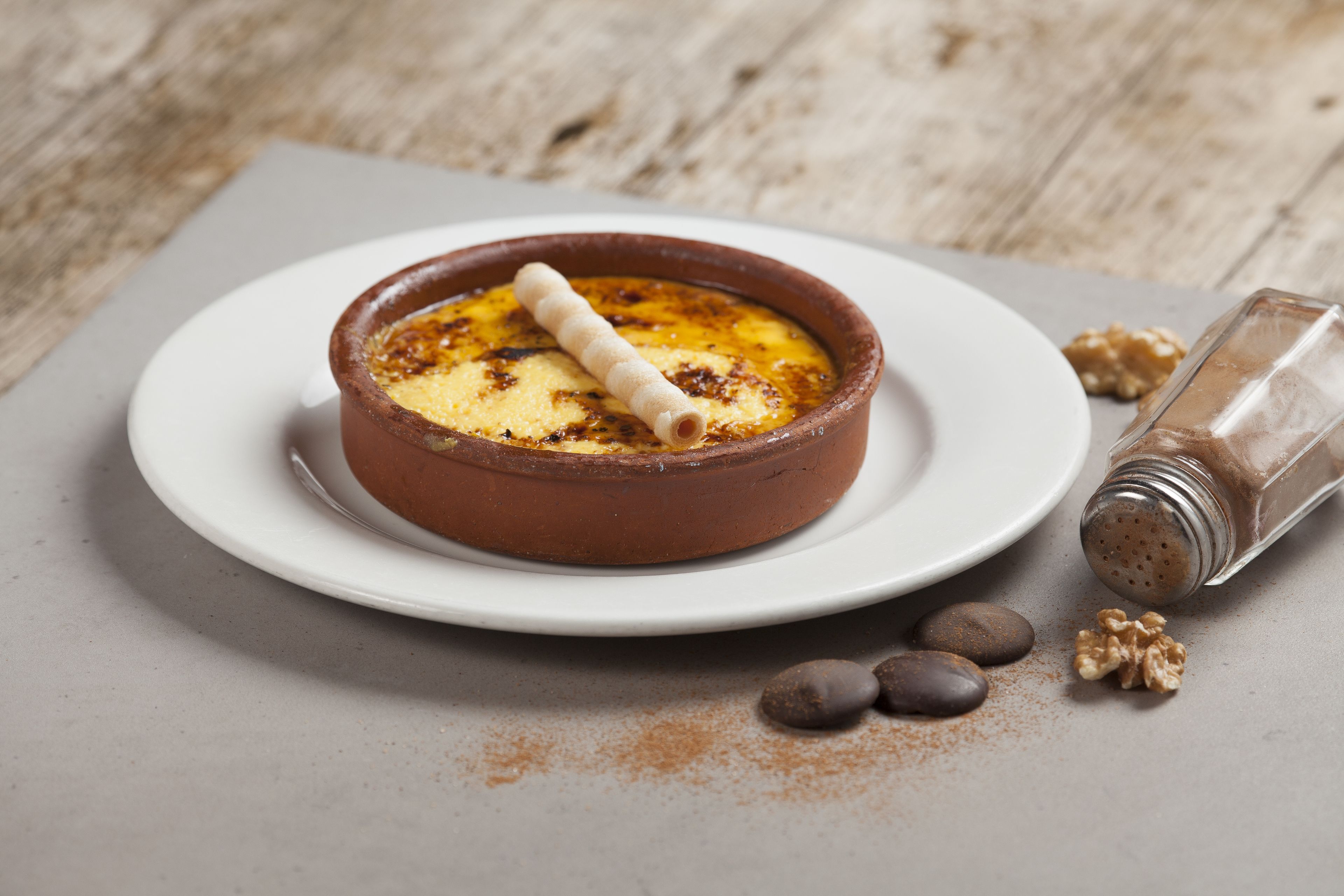 Ein Dessert in einer braunen Kرامیکшапка auf einem weißen Teller, nebenher ein Salzstreuer und Walnüsse.