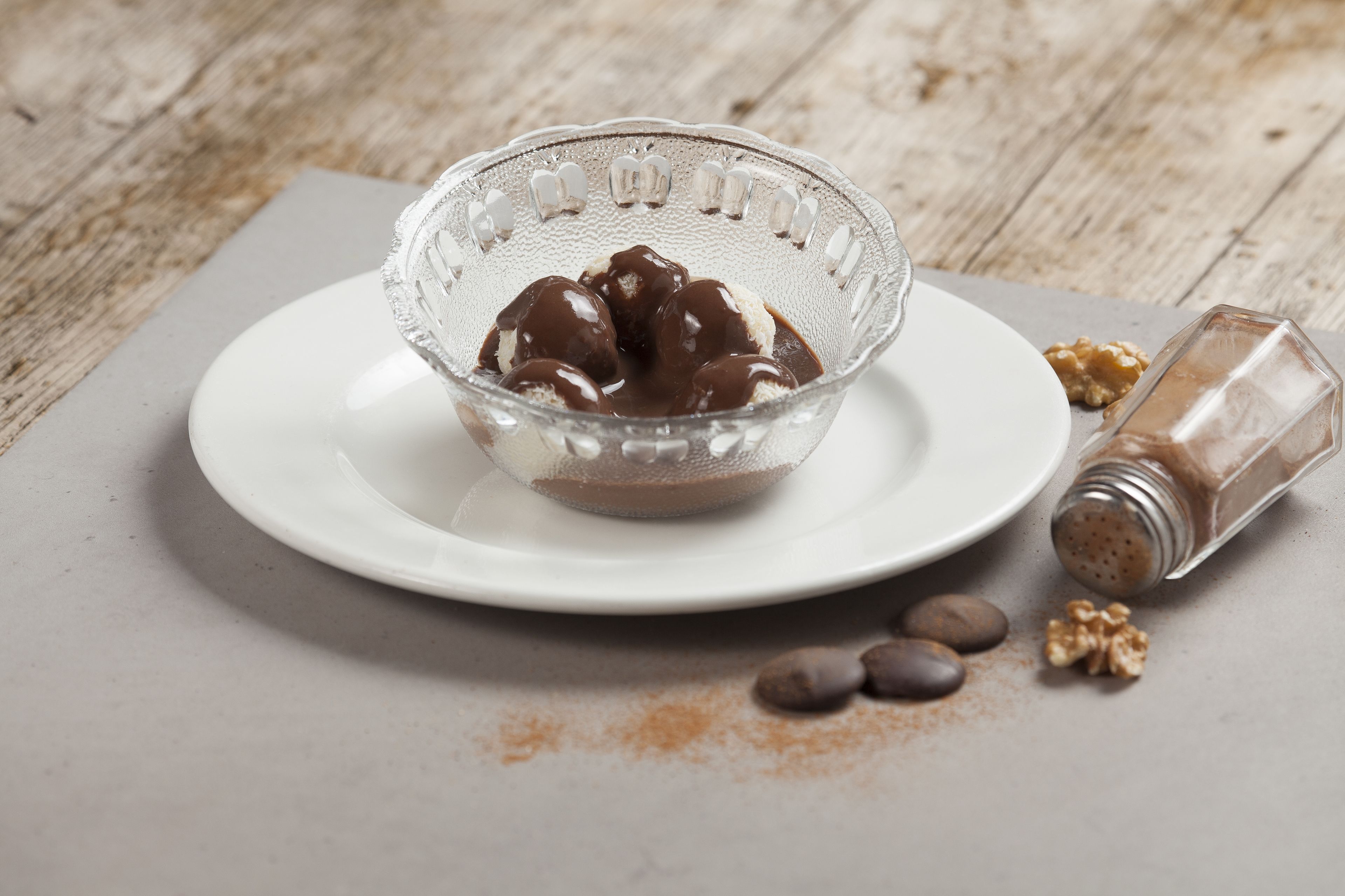 A clear glass bowl with chocolate-covered ice cream on a white plate, accompanied by scattered walnuts and a salt shaker.