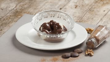 A clear glass bowl with chocolate-covered ice cream on a white plate, accompanied by scattered walnuts and a salt shaker.