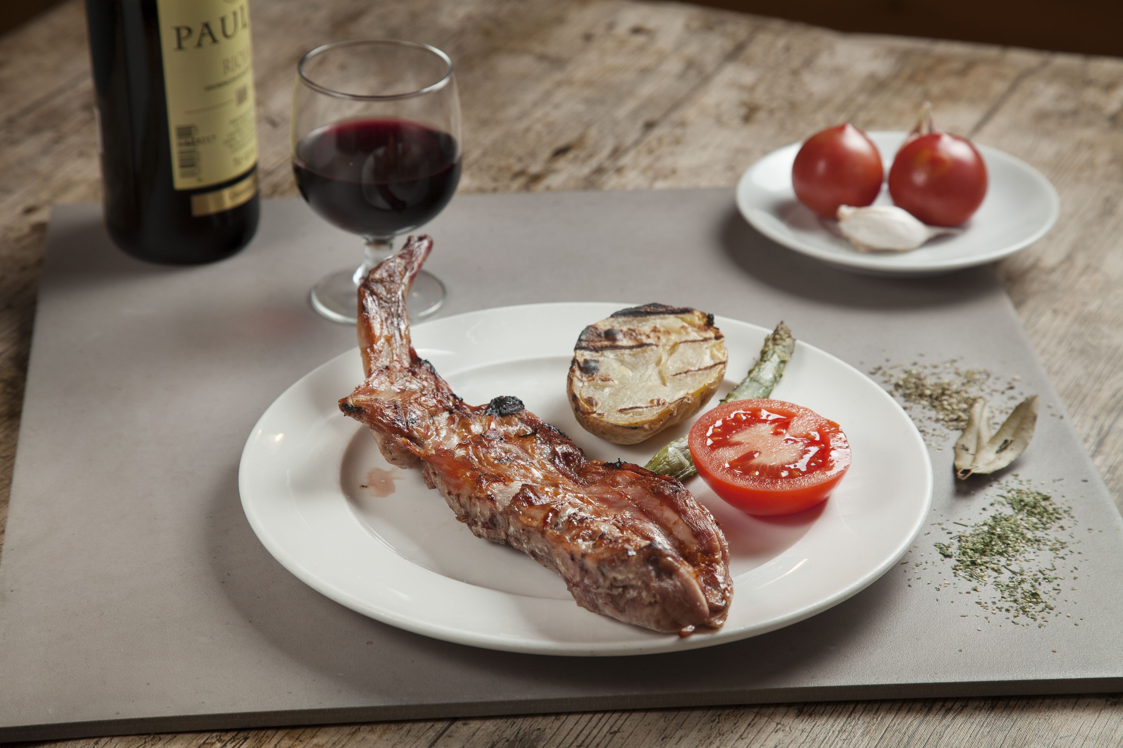 A plate with grilled meat, a grilled tomato, and a roasted potato on a wooden table.