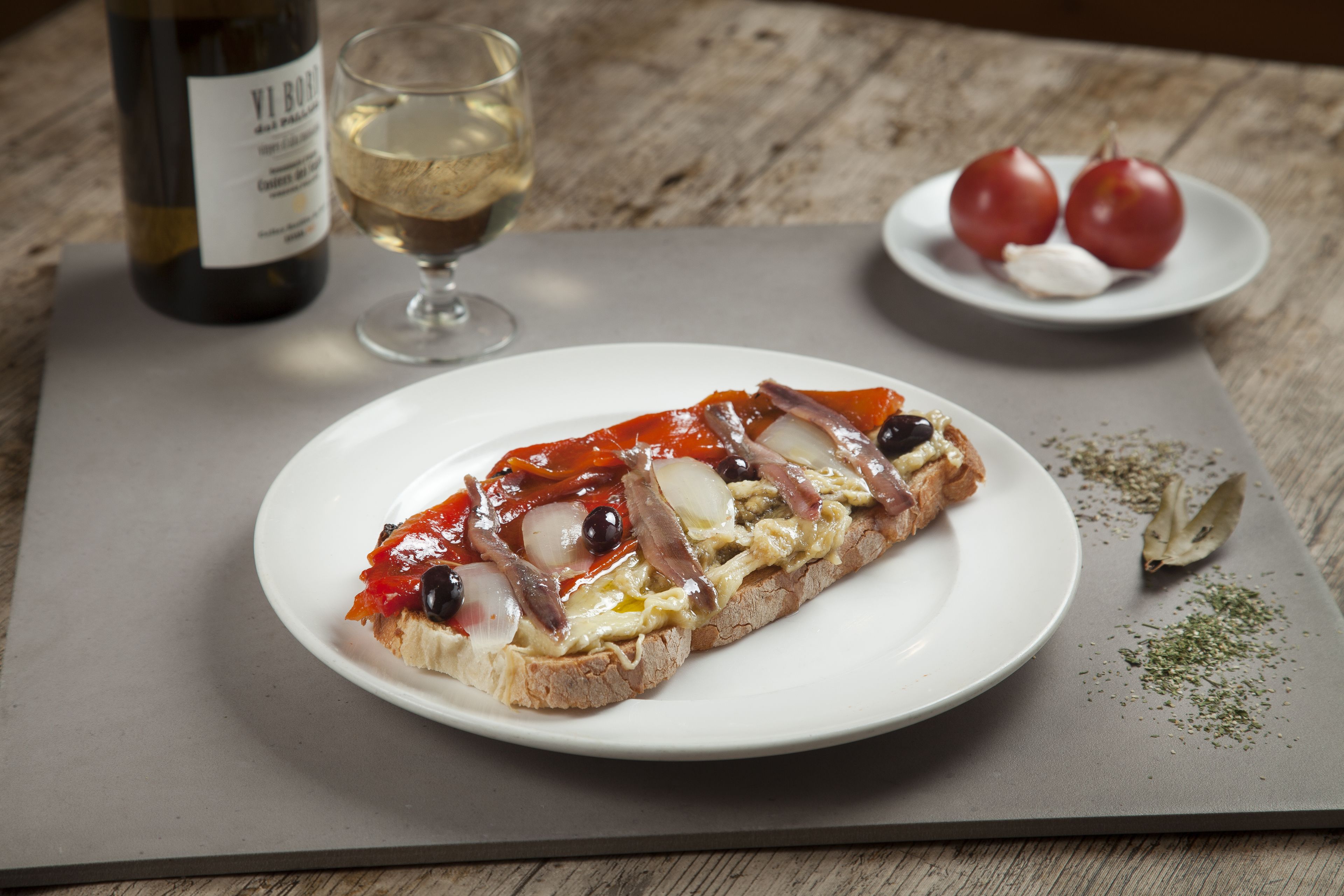 A plate with a sandwich, a glass of white wine, and a plate with tomatoes and garlic on a wooden table.