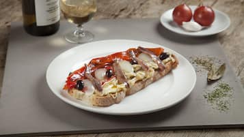 A plate with a sandwich, a glass of white wine, and a plate with tomatoes and garlic on a wooden table.