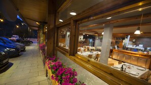 En restaurant med udendørs siddepladser, en parkeringsplads med biler og et vindue med pink blomster.