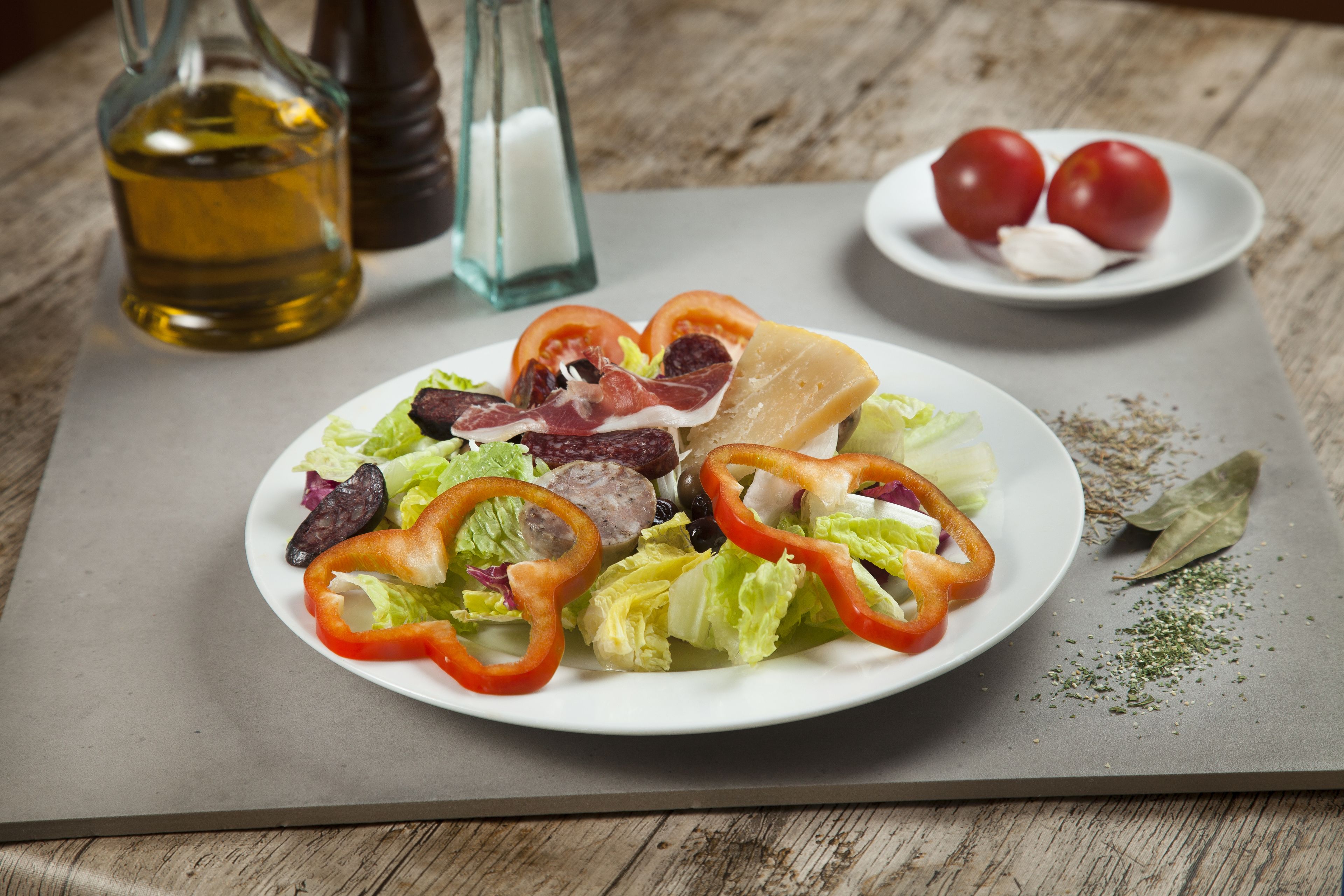 Une assiette de salade composée de divers légumes et de différentes sortes de viande, une petite assiette de tomates cerises et une bouteille d’huile sur une table en bois.