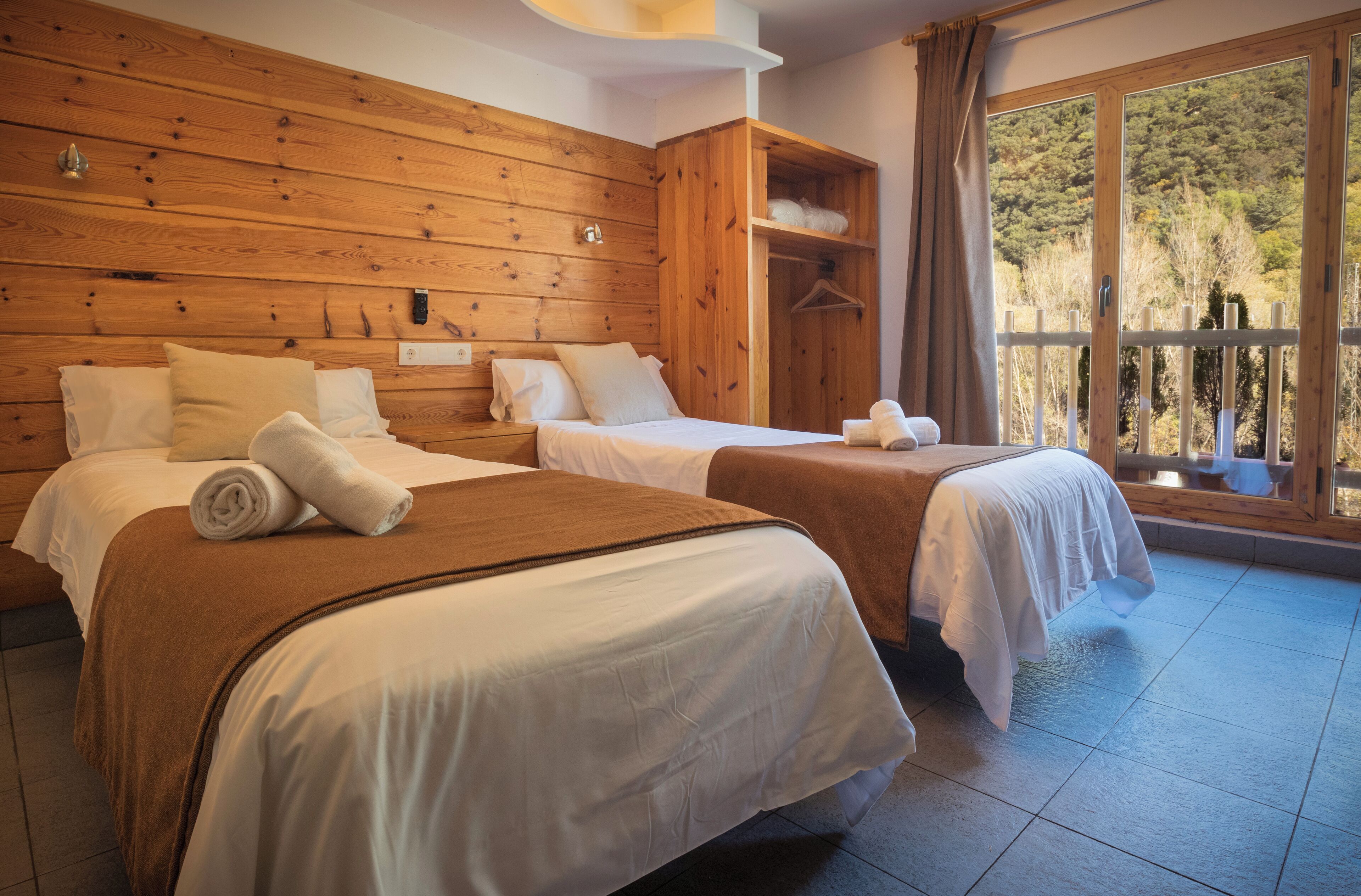 A hotel room with two beds, wooden headboard, large windows with a view of trees, and a balcony.