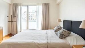 Quarto de hotel com cama, duas almofadas, mesa de cabeceira, luminária e janela com cortinas.