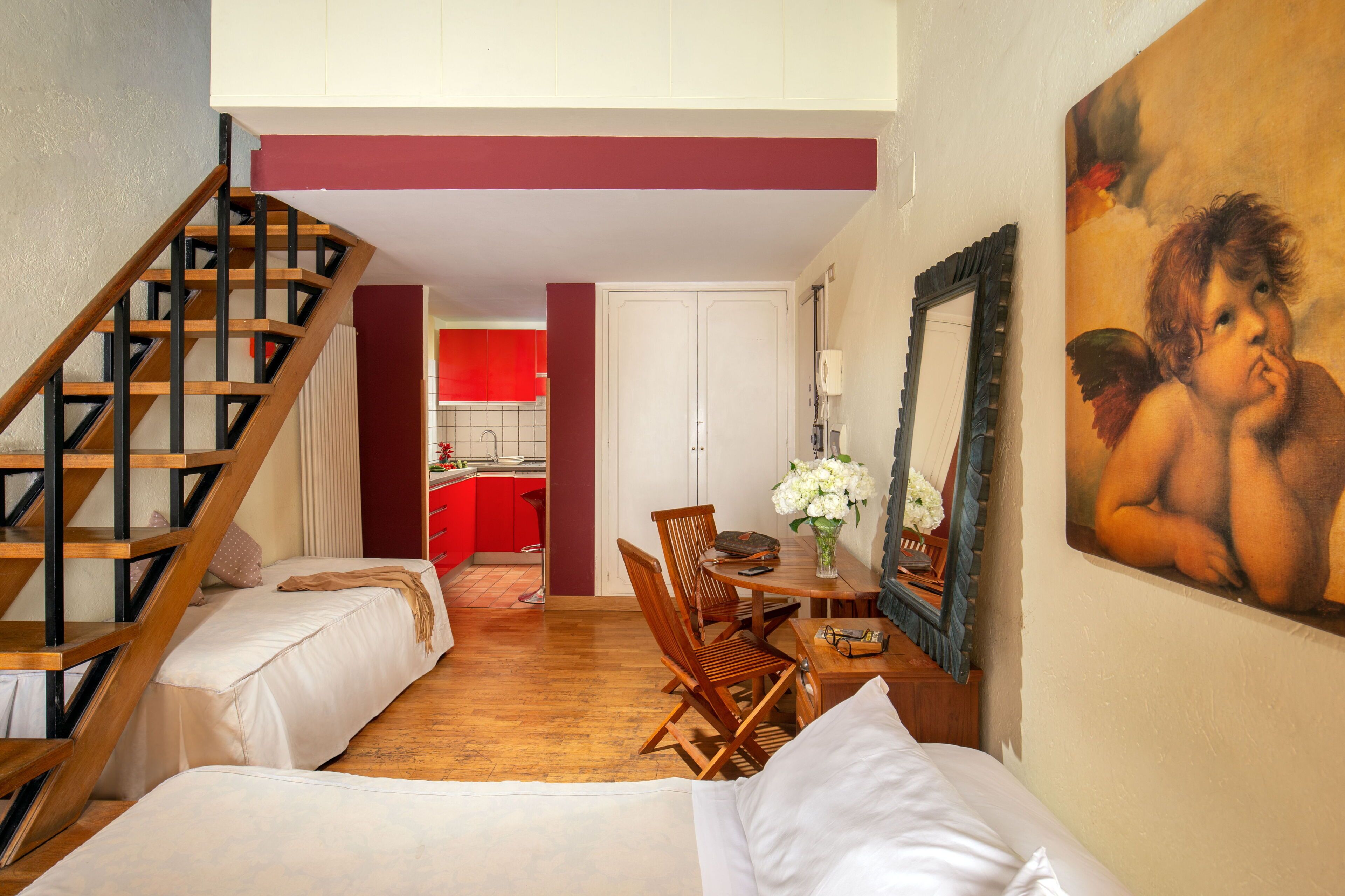 A bedroom with a wooden staircase, a dining area with a red kitchen in the background, and a painting on the wall.