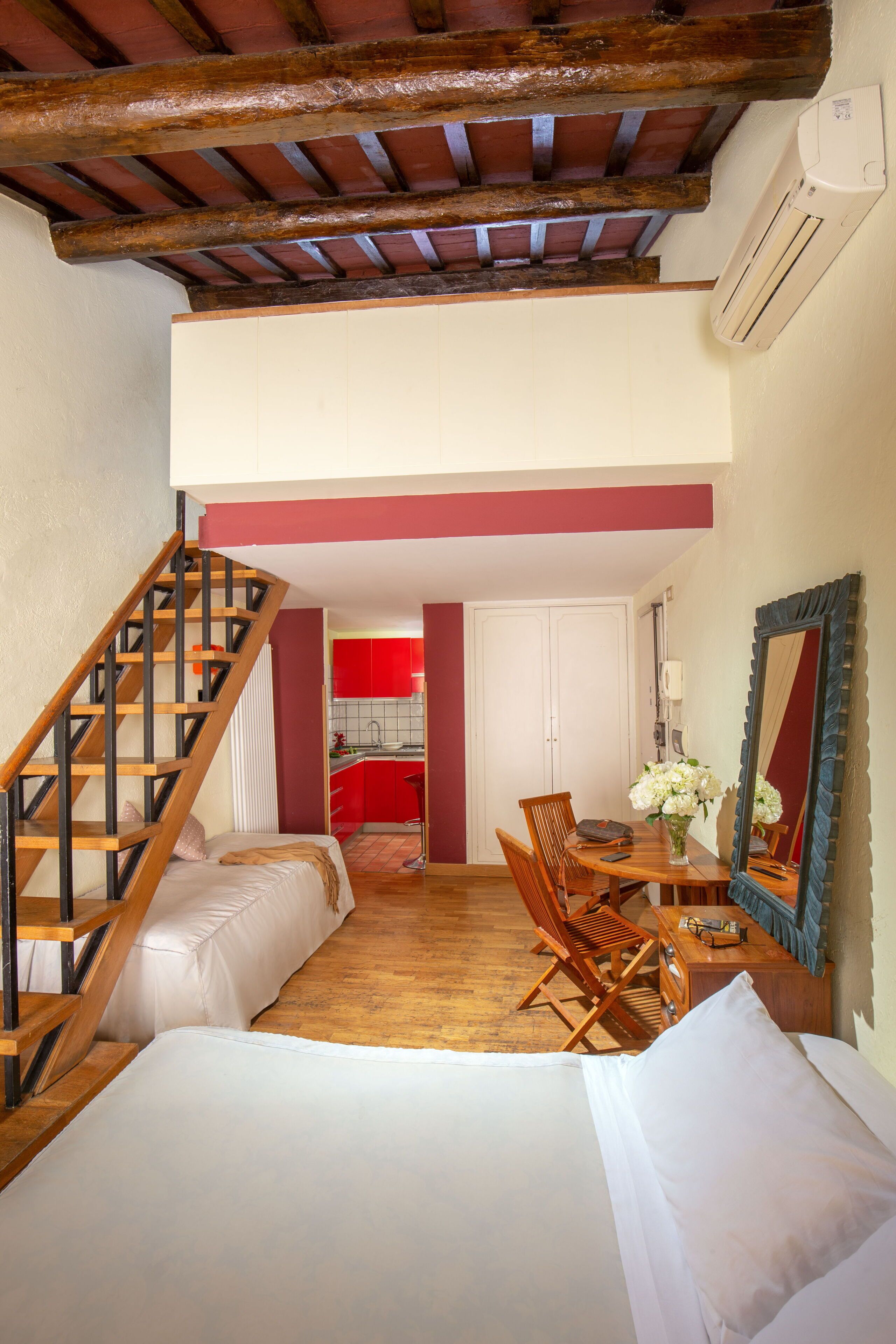 Un dormitorio con una escalera de madera, una zona de comedor con una mesa y sillas, y vista a una cocina con armarios rojos.