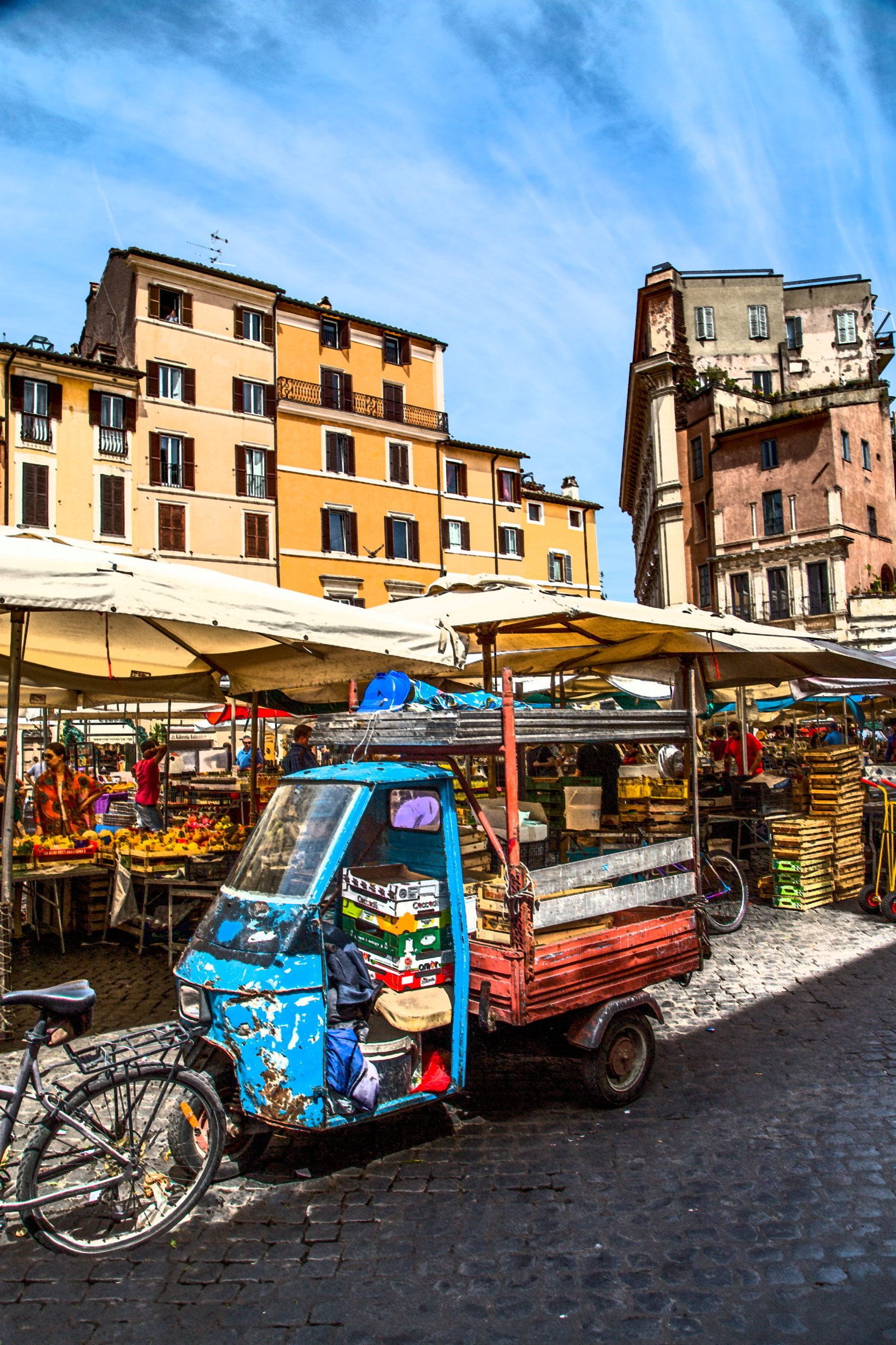 A street market with a blue three-wheeled cart selling fruits and vegetables.