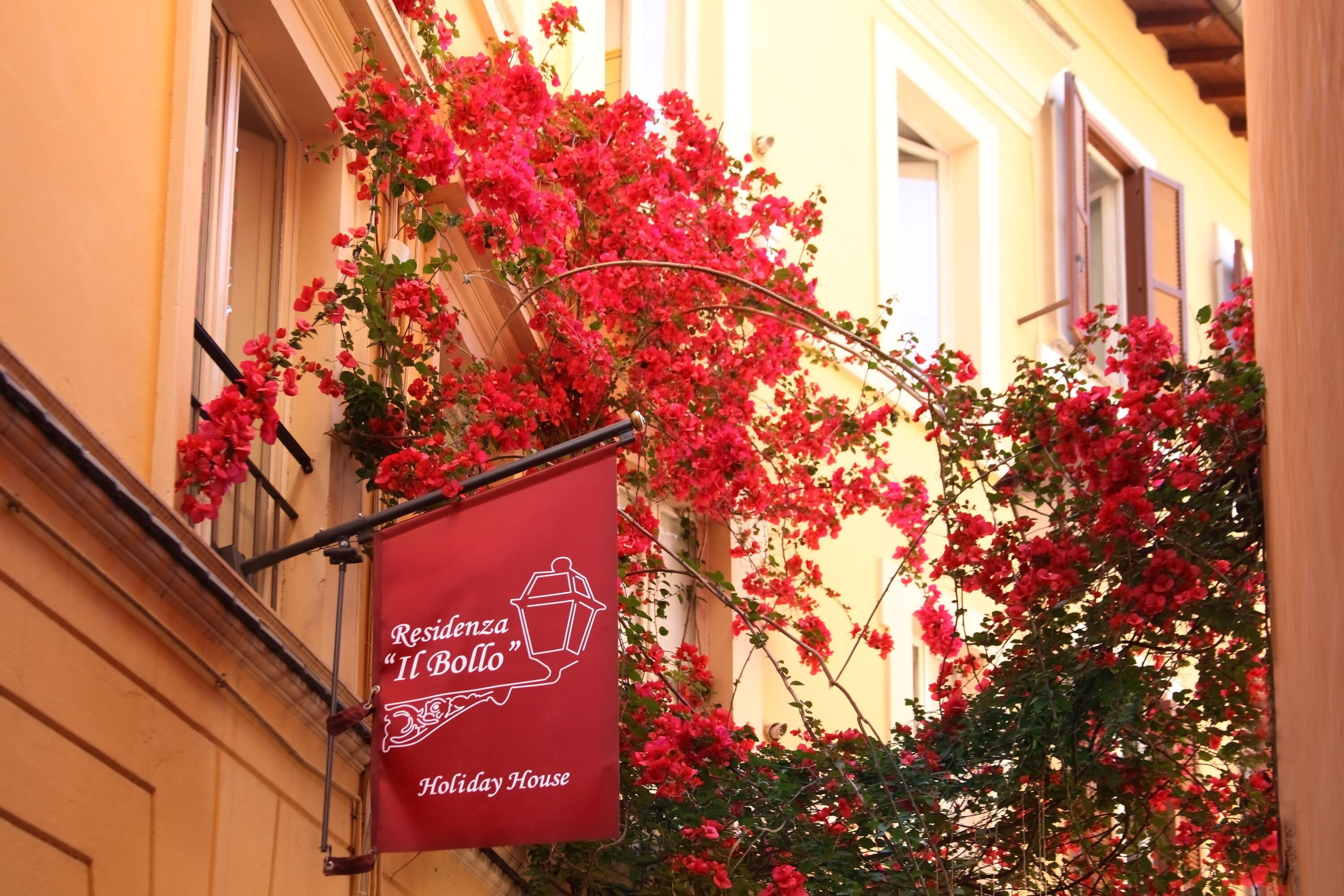 Une banderole rouge portant les inscriptions « Residenza Il Bollo » et « Maison de vacances », placée devant la façade d’un immeuble doté d’un balcon et ornée de plantes à fleurs rouges.