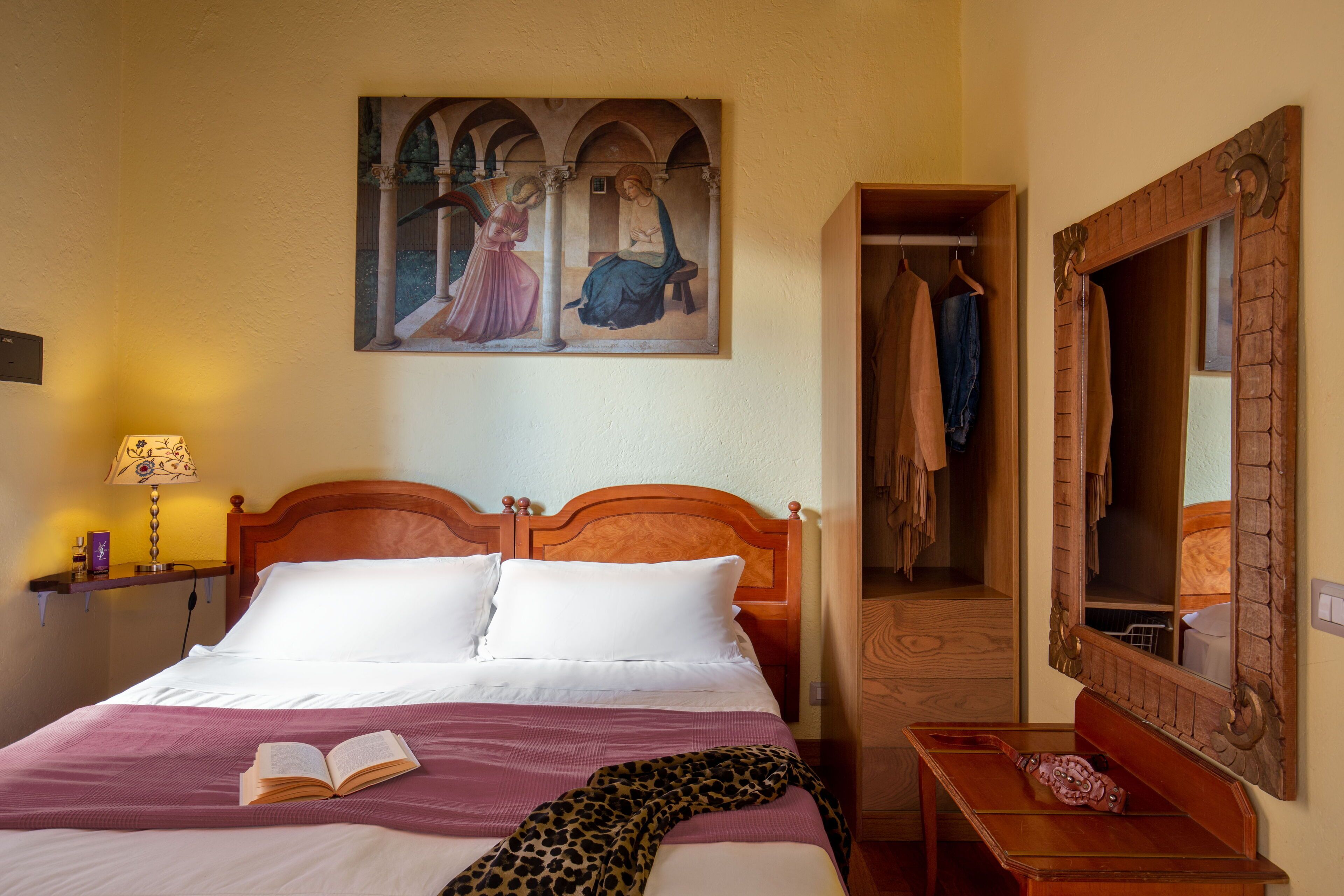 A neatly made bed with a book, a painting above it, and a mirror on the wall.