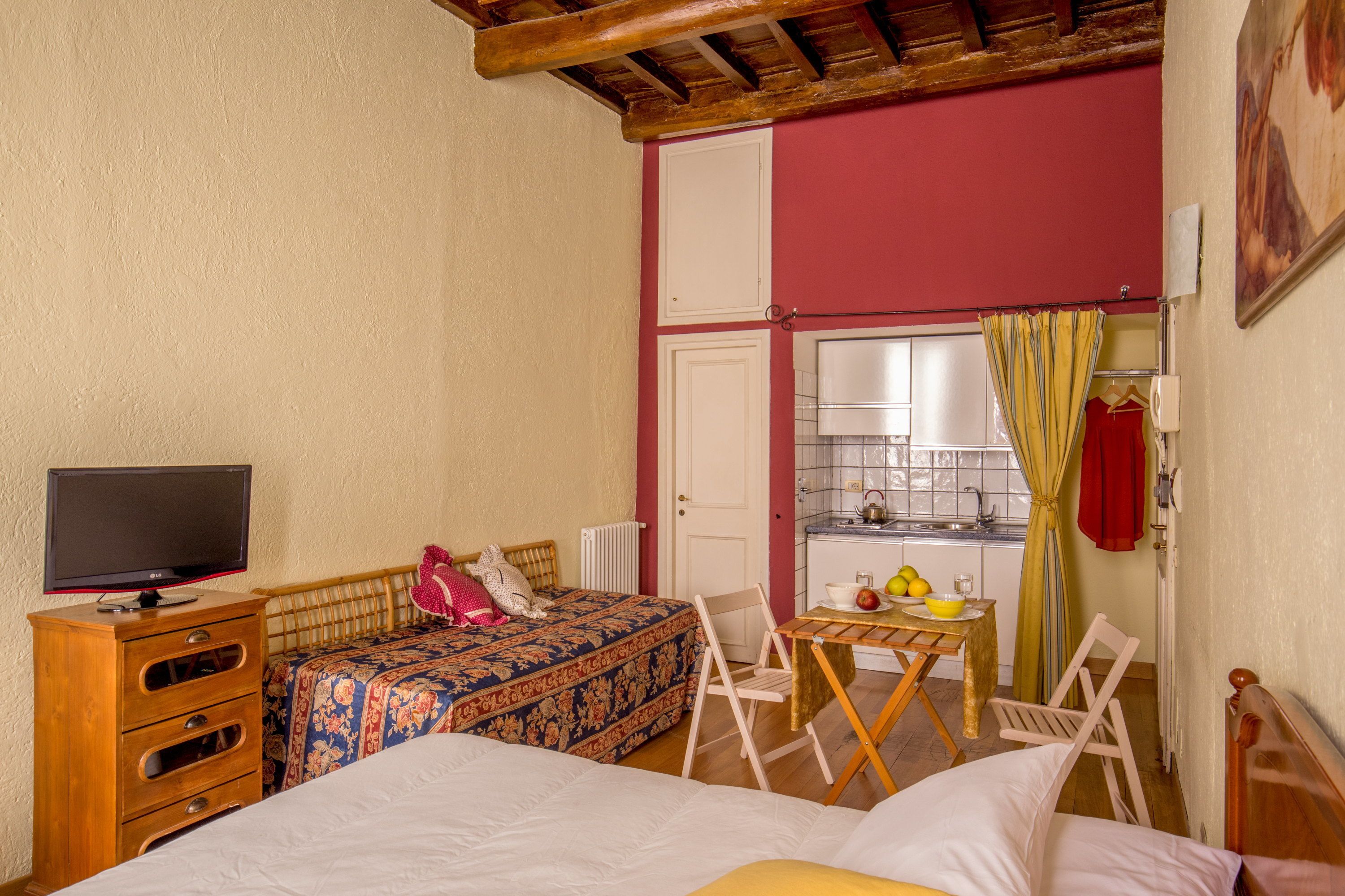 A bedroom with a bed, a TV, a small table with a bowl of fruit, and a window with curtains.