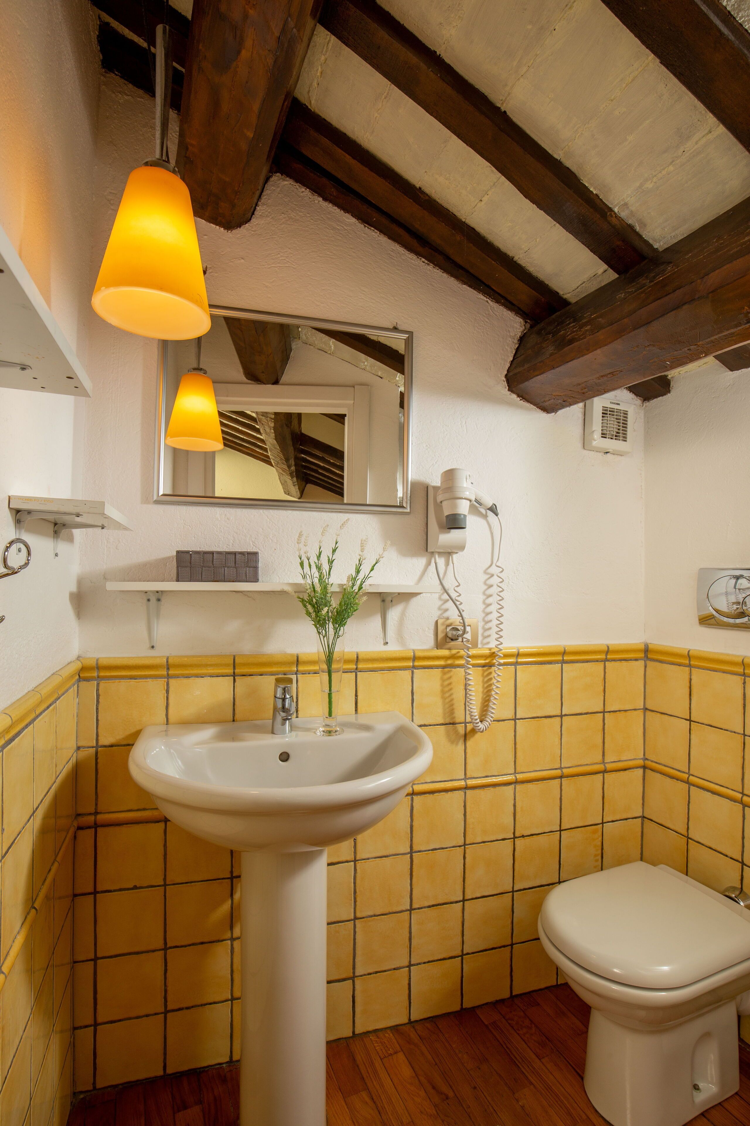 A bathroom with a white sink, a toilet, and a mirror above the sink. There is a yellow-tiled wall and a wooden ceiling beam.