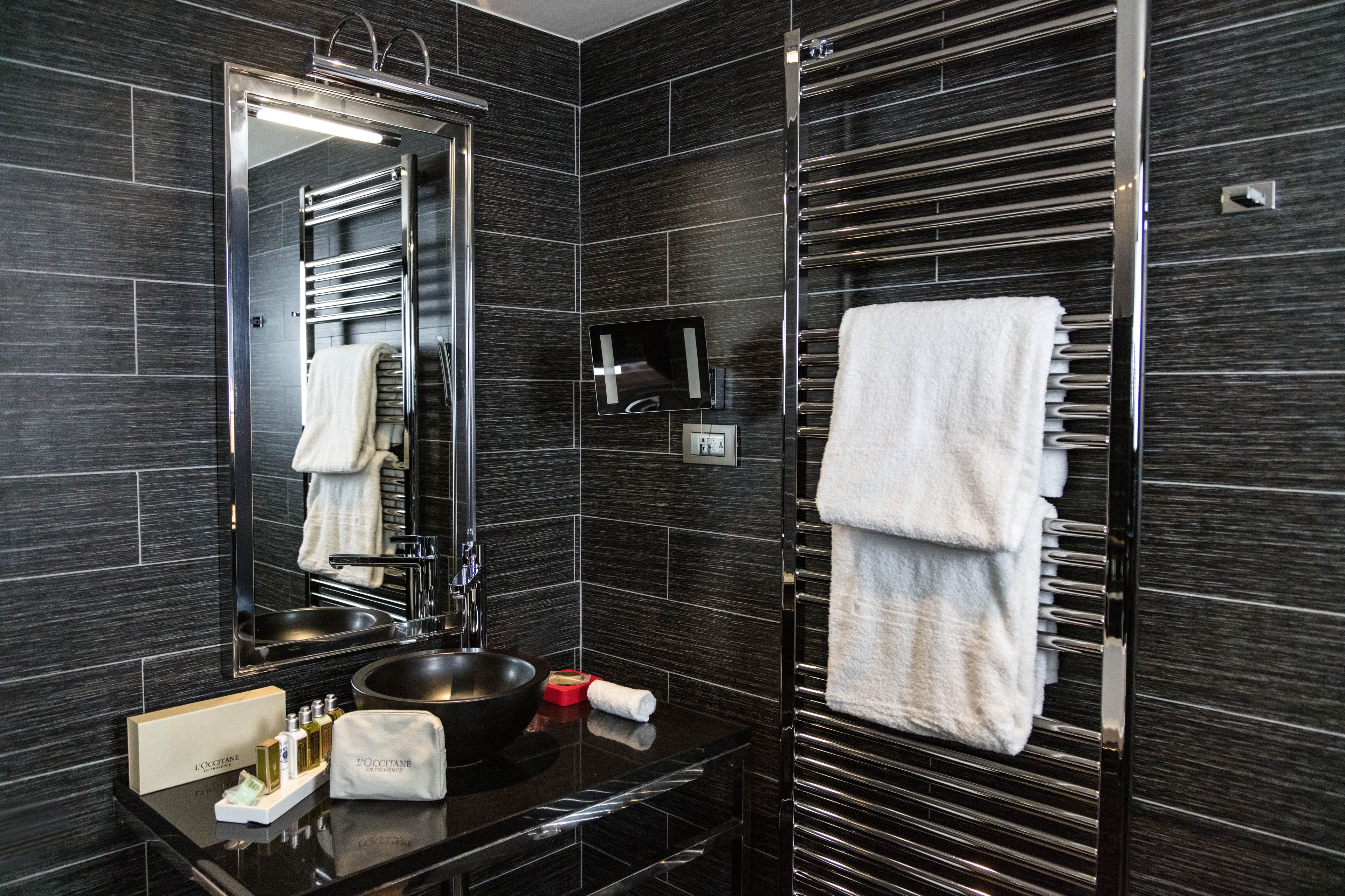 A modern bathroom with dark tiled walls, a large mirror, a black sink, and a towel rack with multiple towels.