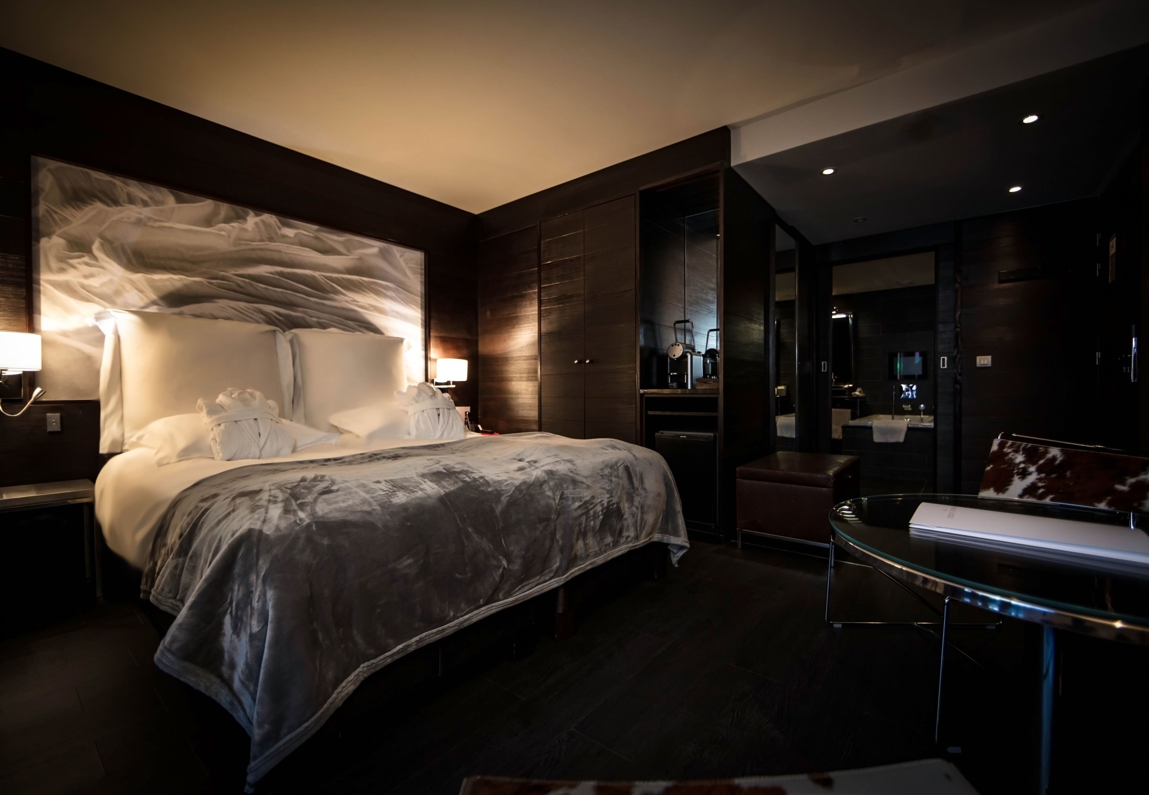 A modern hotel room with a large bed, a glass coffee table, and a dark wood finish.
