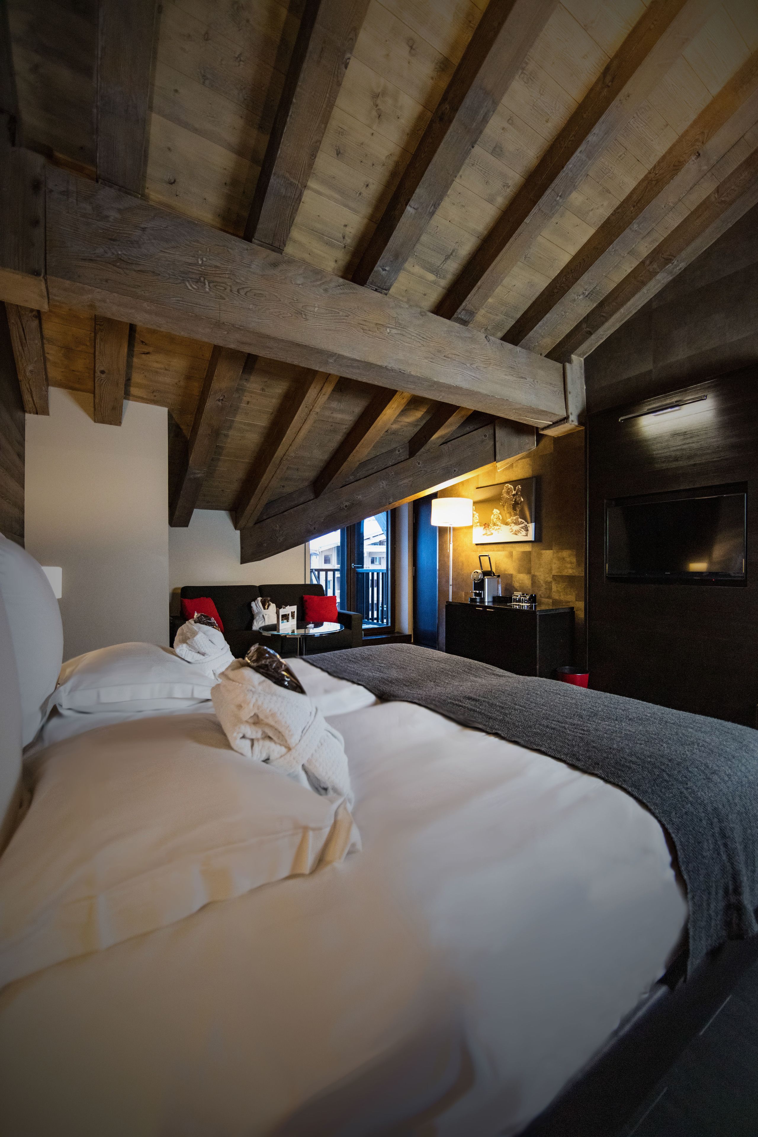 A room with a wooden ceiling, a bed with white linens, a sofa with red cushions, and a desk with a lamp.
