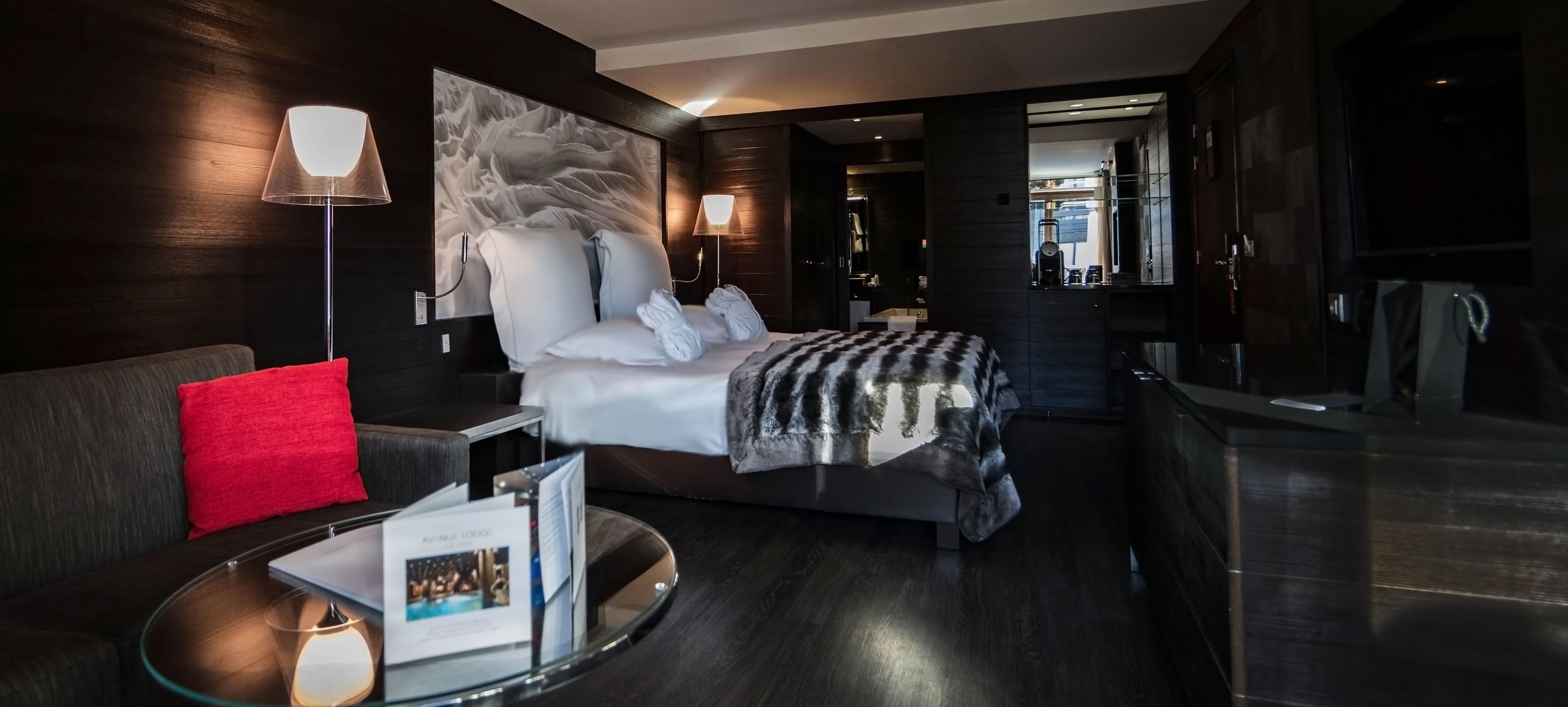 A modern hotel room with a large bed, a glass side table, a red pillow, a wall-mounted lamp, and a framed artwork.