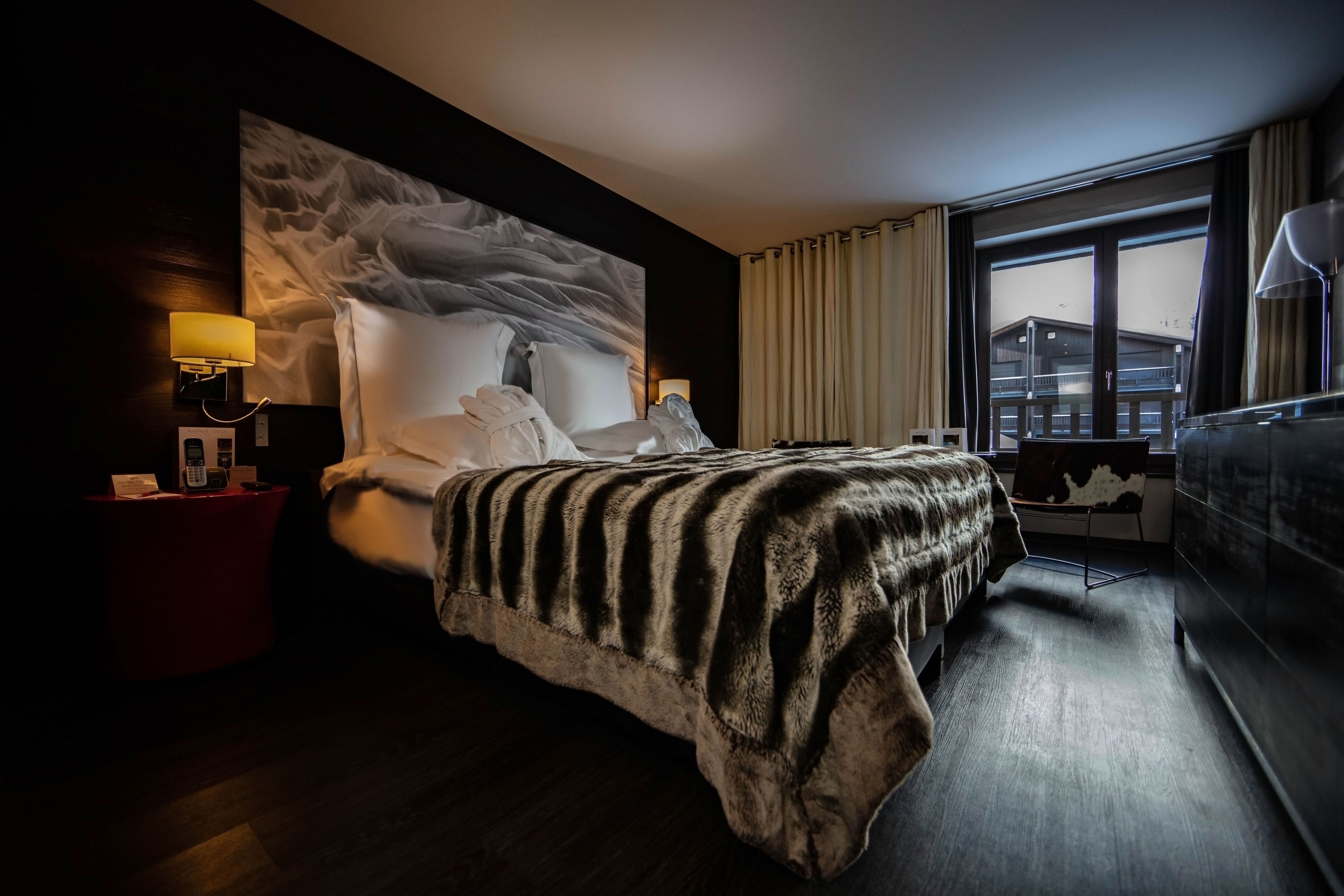 Une chambre d’hôtel avec un grand lit, une table de chevet avec une lampe, une fenêtre avec des rideaux et une vue sur les bâtiments environnants.