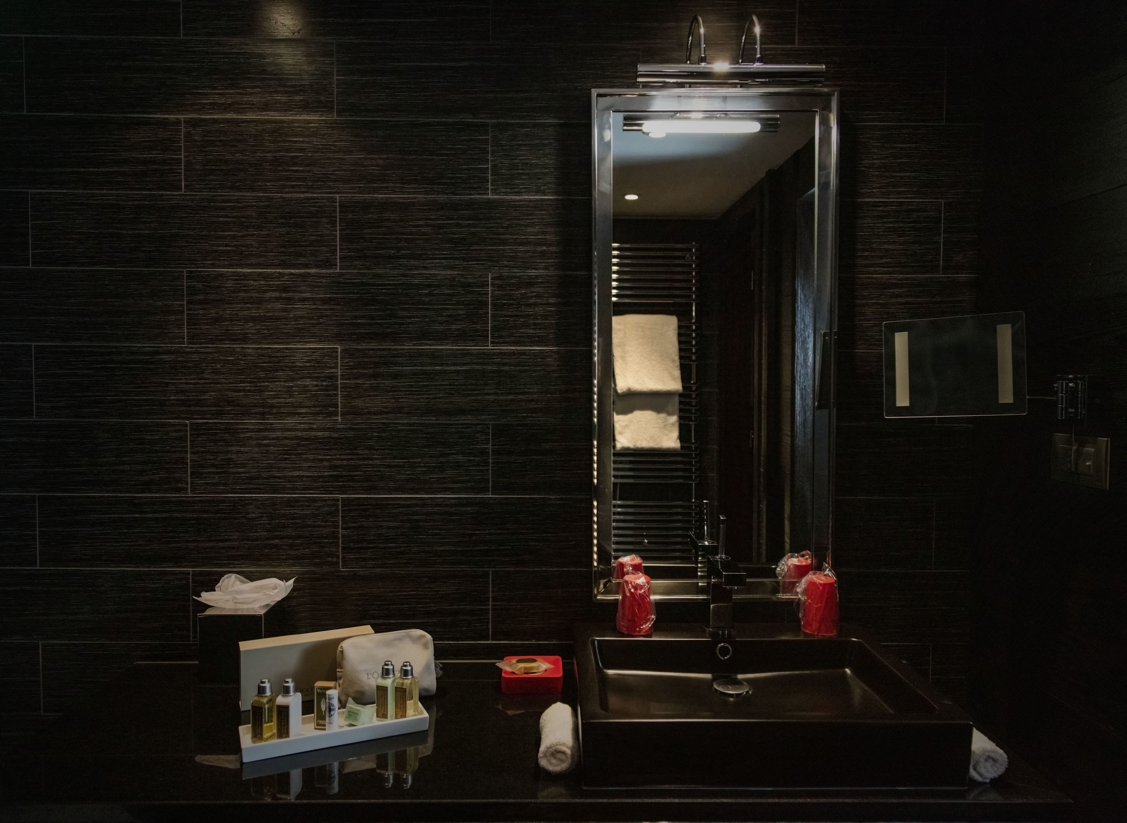 A modern bathroom with a dark tiled wall, a sink with a mirror, and toiletries on the counter.