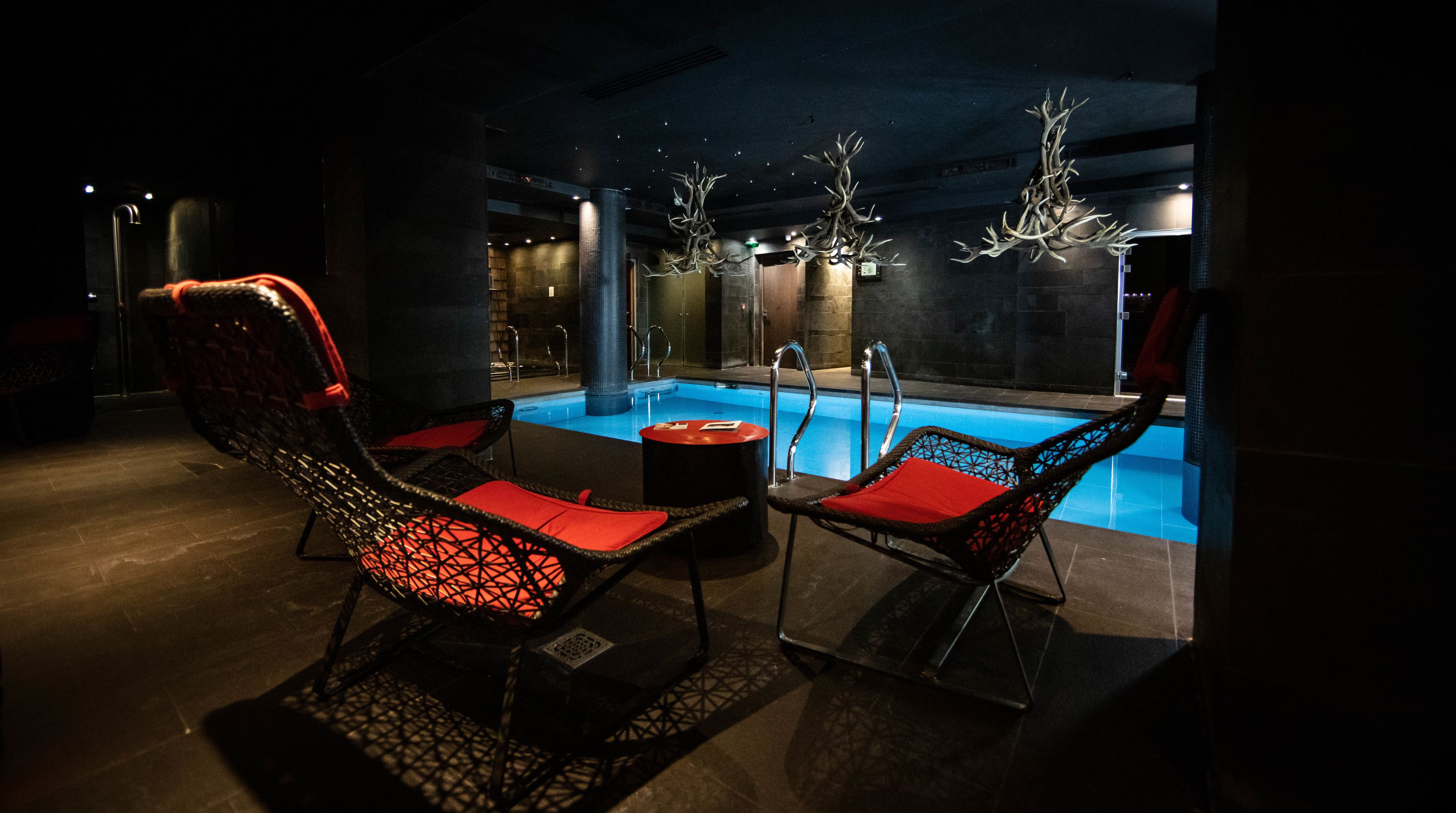 Et svagt oplyst poolområde indendørs med to fletstole med røde hynder, et lille rundt bord og en pool med klar, blå farve.