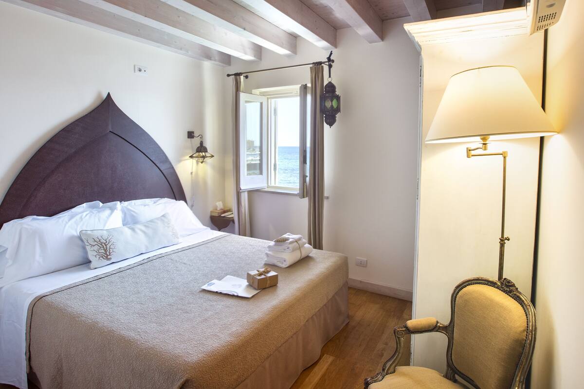 A neatly made bed with a headboard, a bedside table with a lamp, and a view of the sea through an open window.
