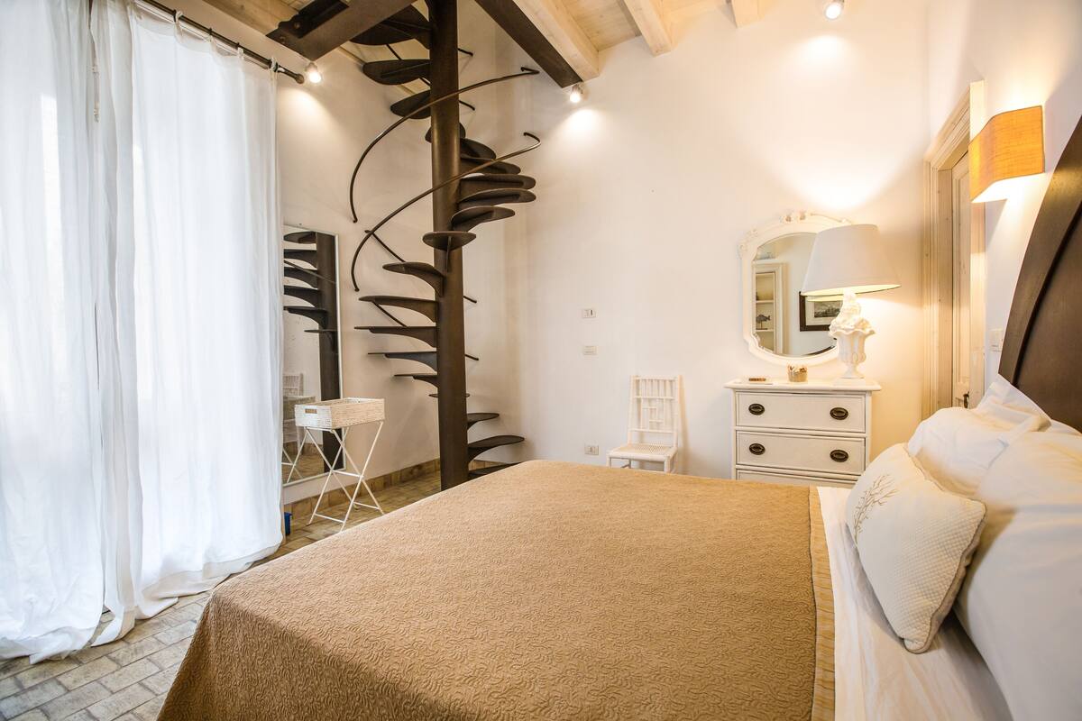 A bedroom with a bed, a nightstand, a chair, a mirror, and a staircase.