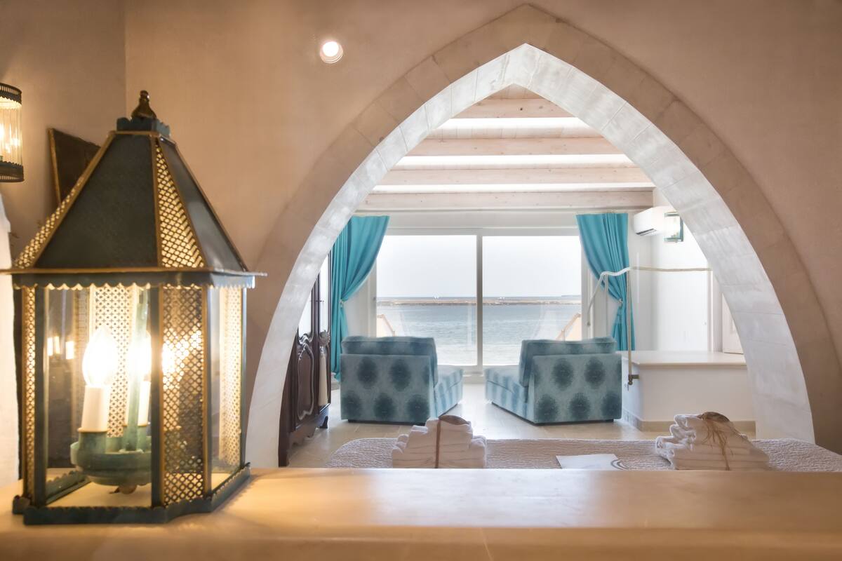 A cozy room with a view of the sea, featuring a large arched doorway, a lantern, and patterned blue sofas.