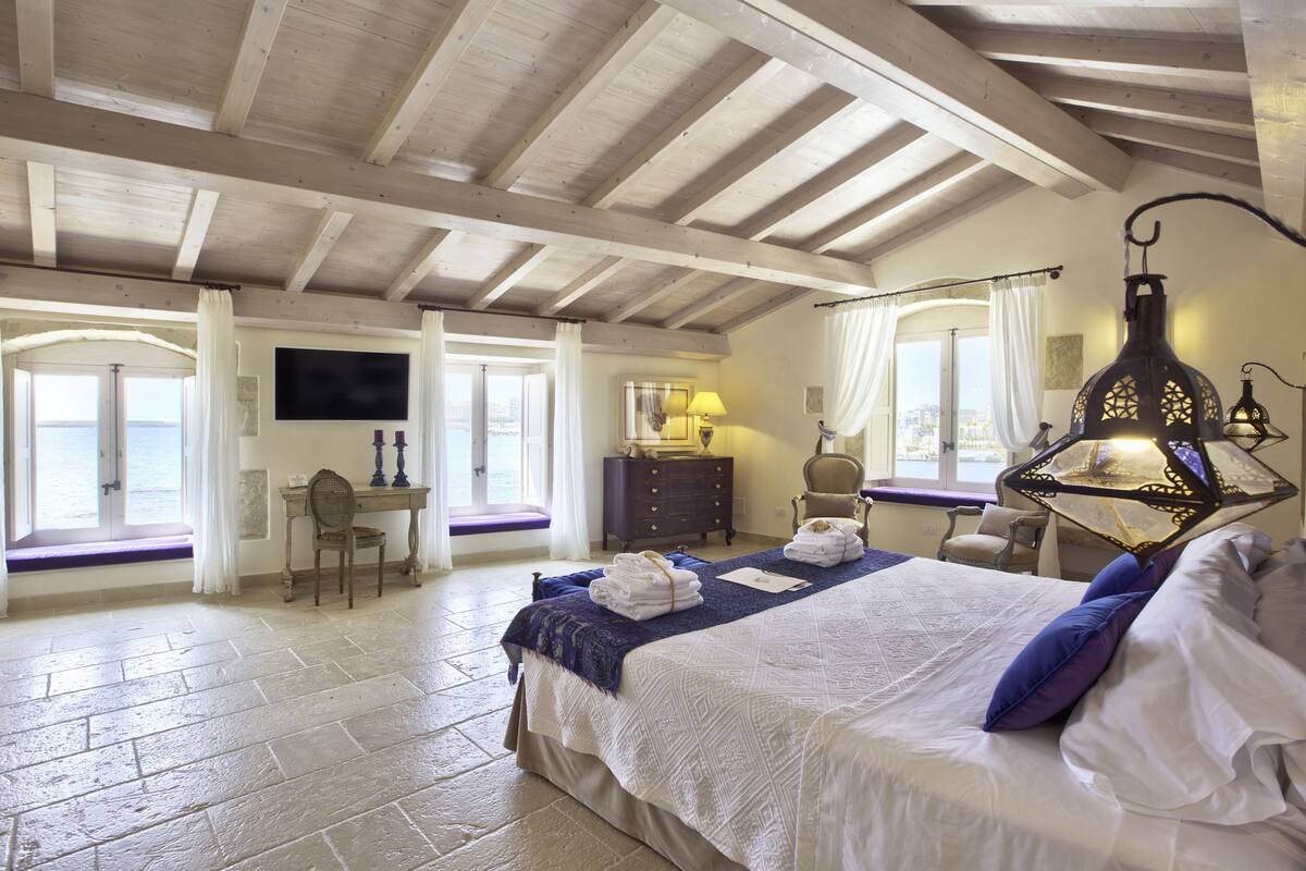A bedroom with a large bed, a desk, a chair, and a TV. There are windows with curtains, a lamp, and a decorative lantern.