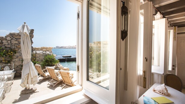 Una habitación con un ventanal grande que da a la costa, una mesa con un libro y una silla.