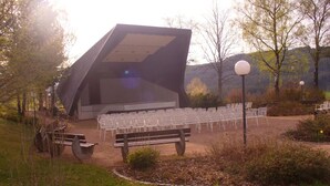 An outdoor amphitheater with a modern design, surrounded by chairs and benches, set in a natural landscape with trees and hills in the background.
