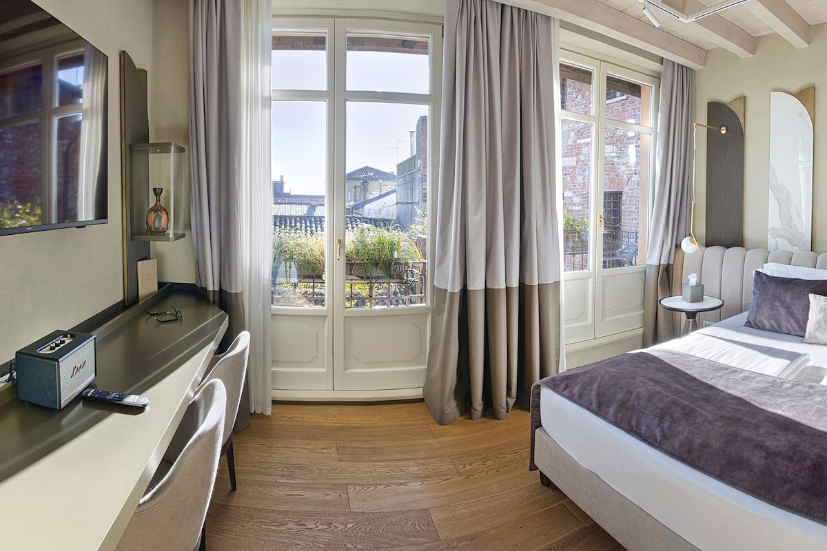 A hotel room with a bed, bedside tables, a desk with a computer, a TV, and a view of the city through large windows.