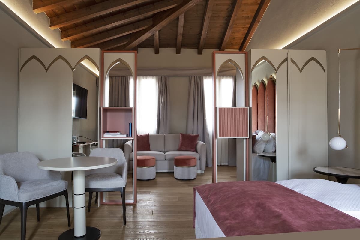 A modern hotel room with a wooden ceiling, a bed with a red blanket, a round table, two armchairs, a sofa, and a TV.