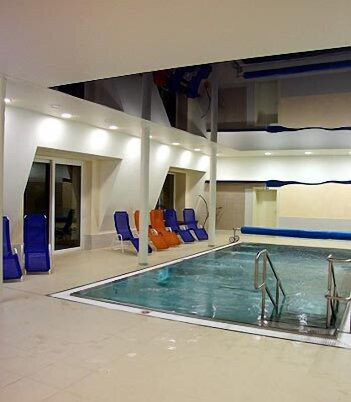 Ein Hallenbad mit blauen und orangefarbenen Liegestühlen, einer Poolleiter und einer Decke mit wellenförmigem Design.